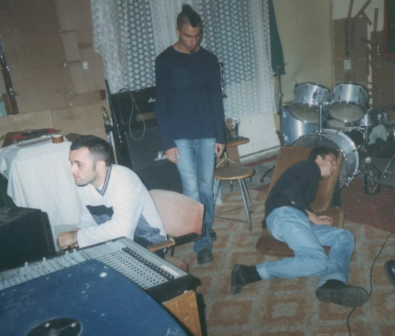 Three men in a music rehearsal room with musical equipment. One man is sitting at a sound mixing console, another is standing with eyes closed, and the third is lying on the floor. The room has musical instruments and amplifiers.