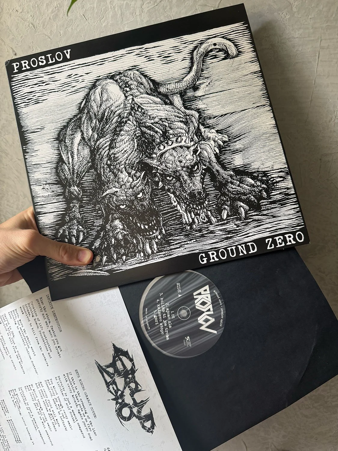 Black and white album cover with a detailed illustration of a four-legged creature with a wolf-like head, muscular body, and tail, accompanied by two snarling wolf heads; top left labeled 'PROSLOV', bottom right labeled 'GROUND ZERO'.