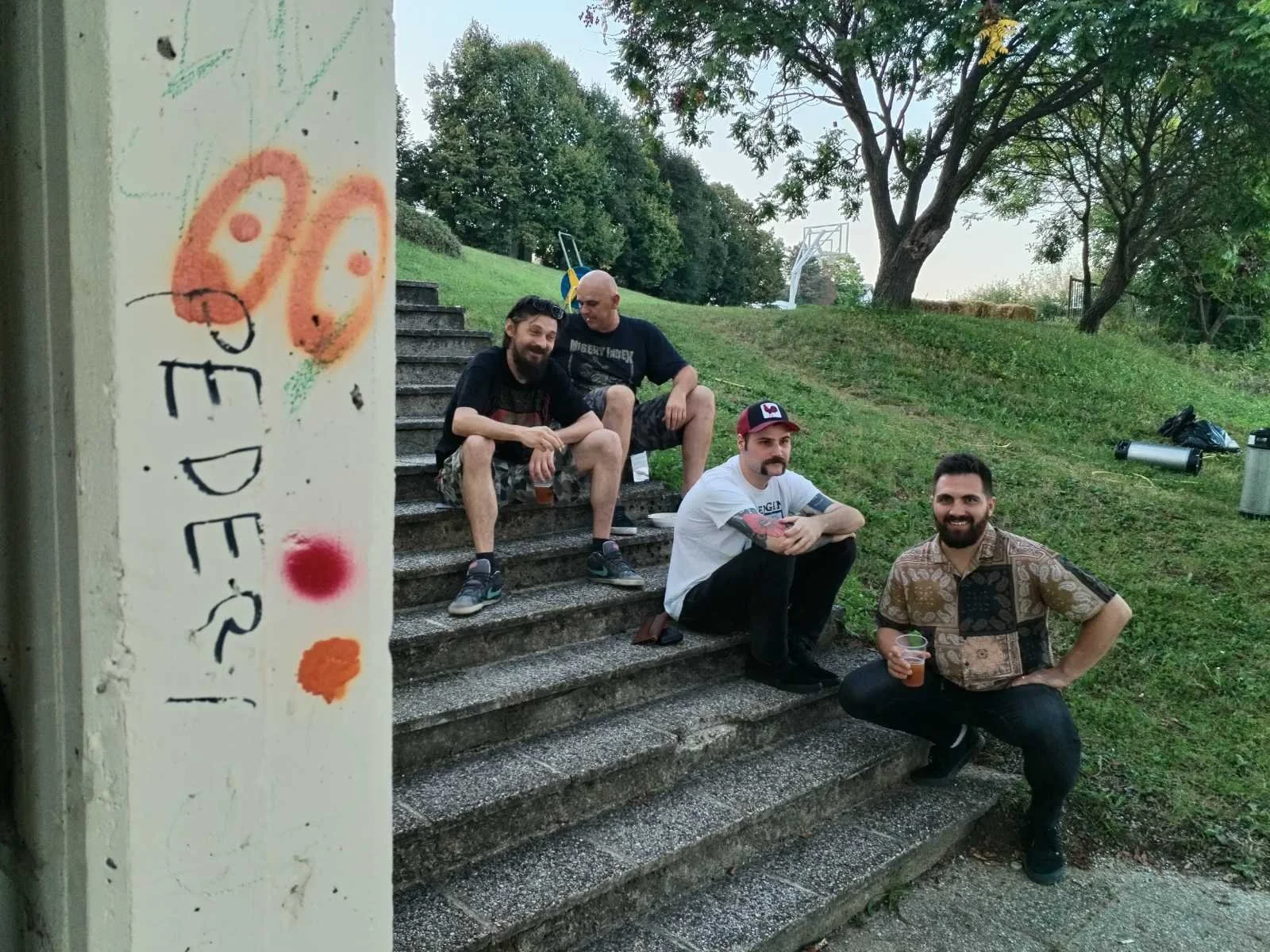 Four men sitting on outdoor concrete stairs in a park, smiling and holding drinks, with trees and grassy area in the background, and some trash and a basketball hoop visible.