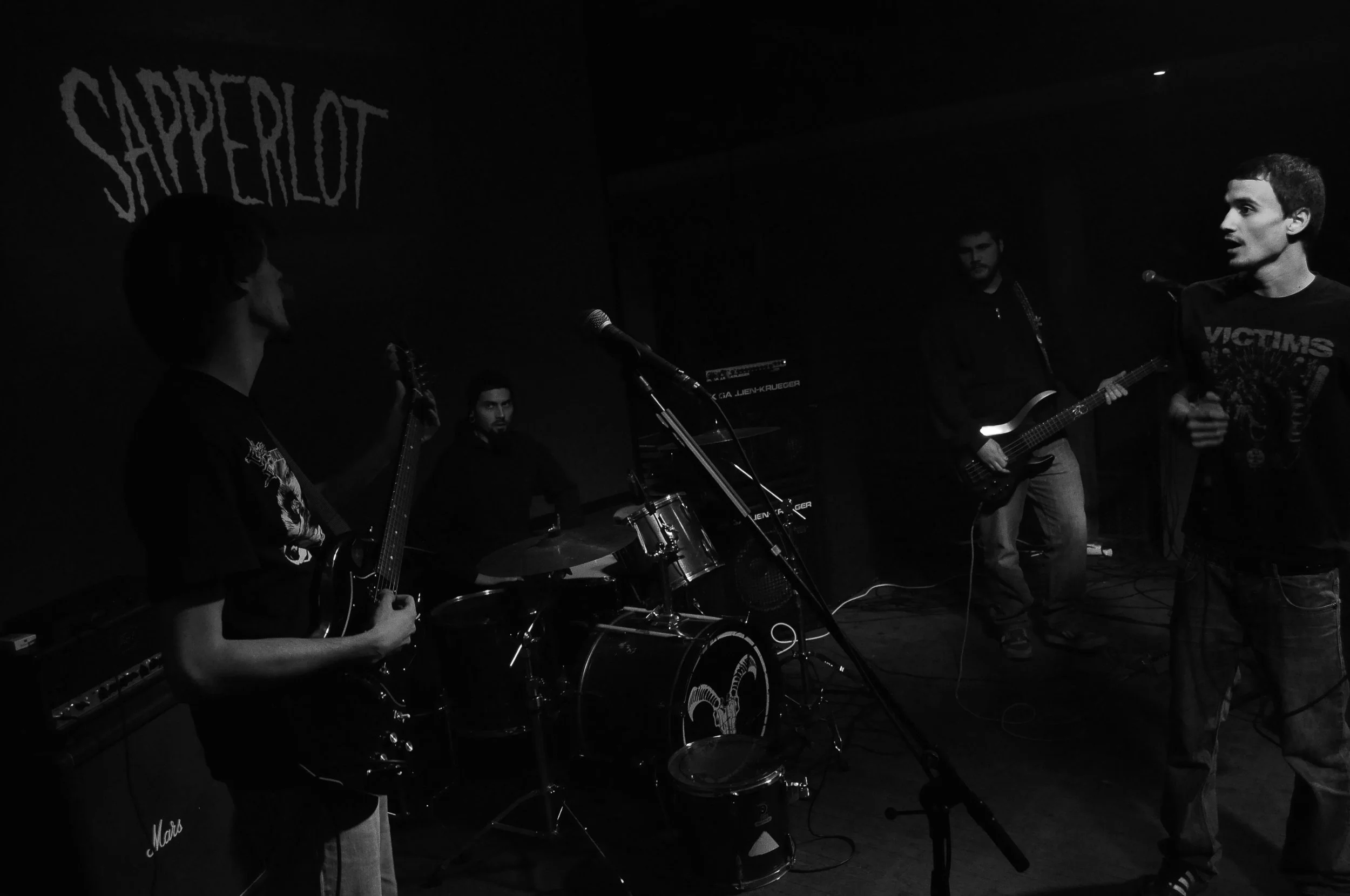 A band performing on stage with four male members, one playing bass, one on drums, one singing, and another with a guitar, in a dark venue with a "Sapperlot" sign in the background.