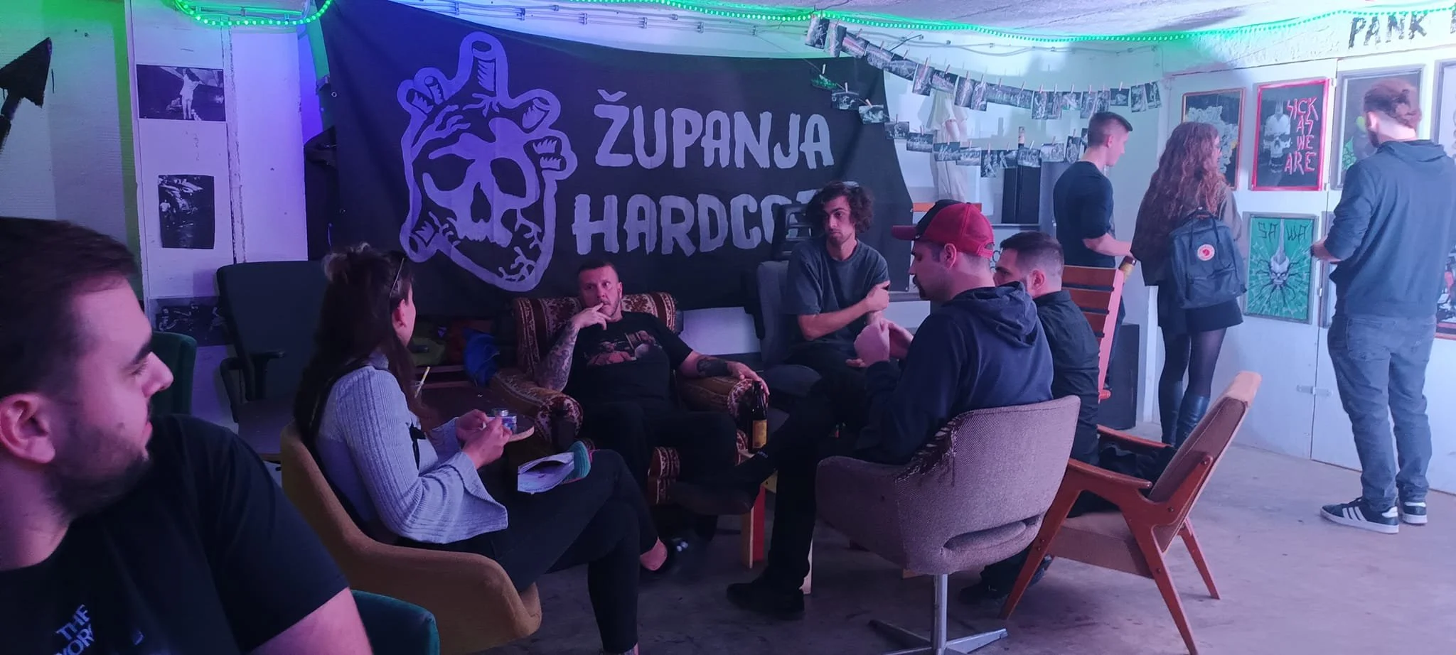 Group of people gathering in a room decorated with neon lights, artwork, and photos on the walls, with a black banner that reads 'ZUPANJA HARDCORE' featuring a stylized heart with a skull inside.