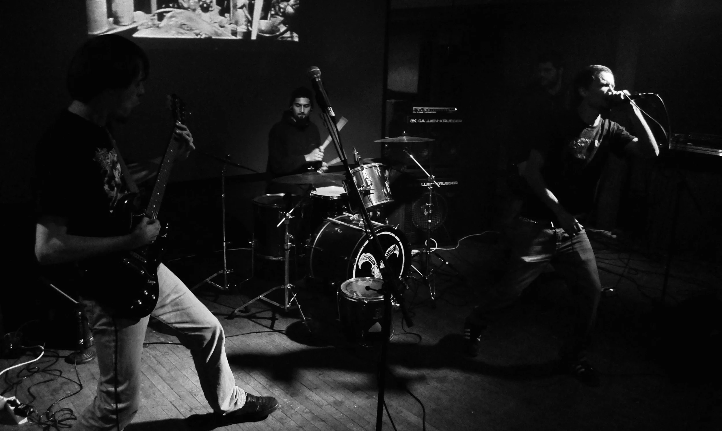 Black and white photo of a band performing on stage. Three members are visible: one playing an electric guitar on the left, another on drums in the center, and a third singer with a microphone on the right.