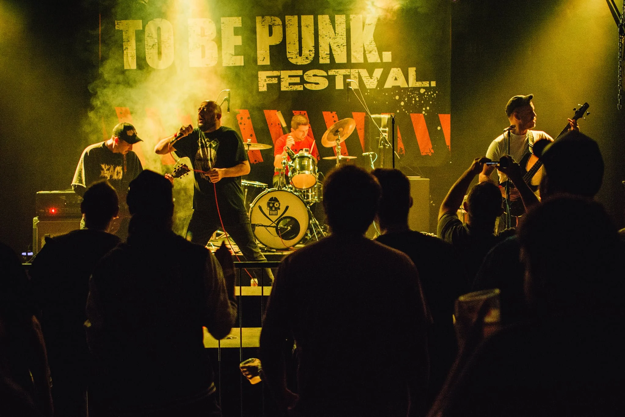 A band performs on stage at a music festival called 'TO BE PUNK FESTIVAL,' with five members playing instruments and a singer holding a microphone in front of an audience.