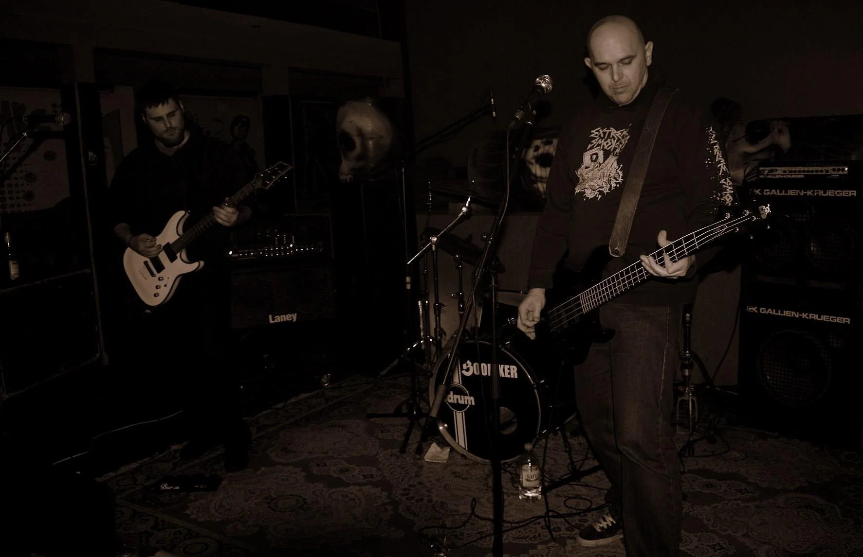 A band performing on stage with two guitarists and a drummer. The setting appears dark with musical equipment visible, including amplifiers and drums.