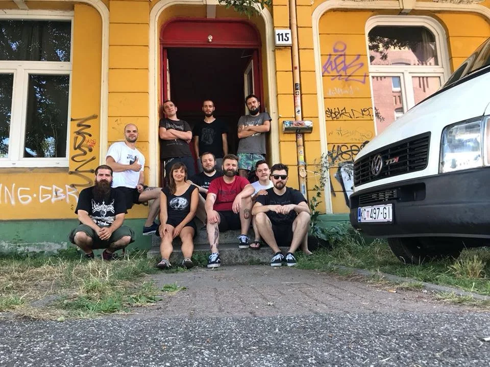 A group of eleven people sitting and standing on the steps of a yellow building with graffiti, next to a white van with a European license plate.