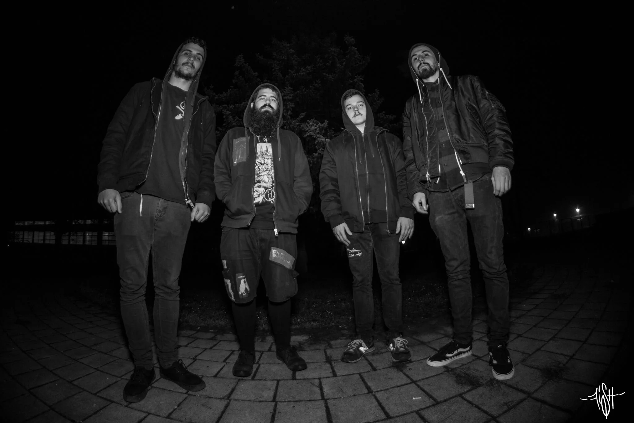 Four young men standing outside at night on a cobblestone path, wearing hoodies and jackets, with a dark sky and trees behind them.