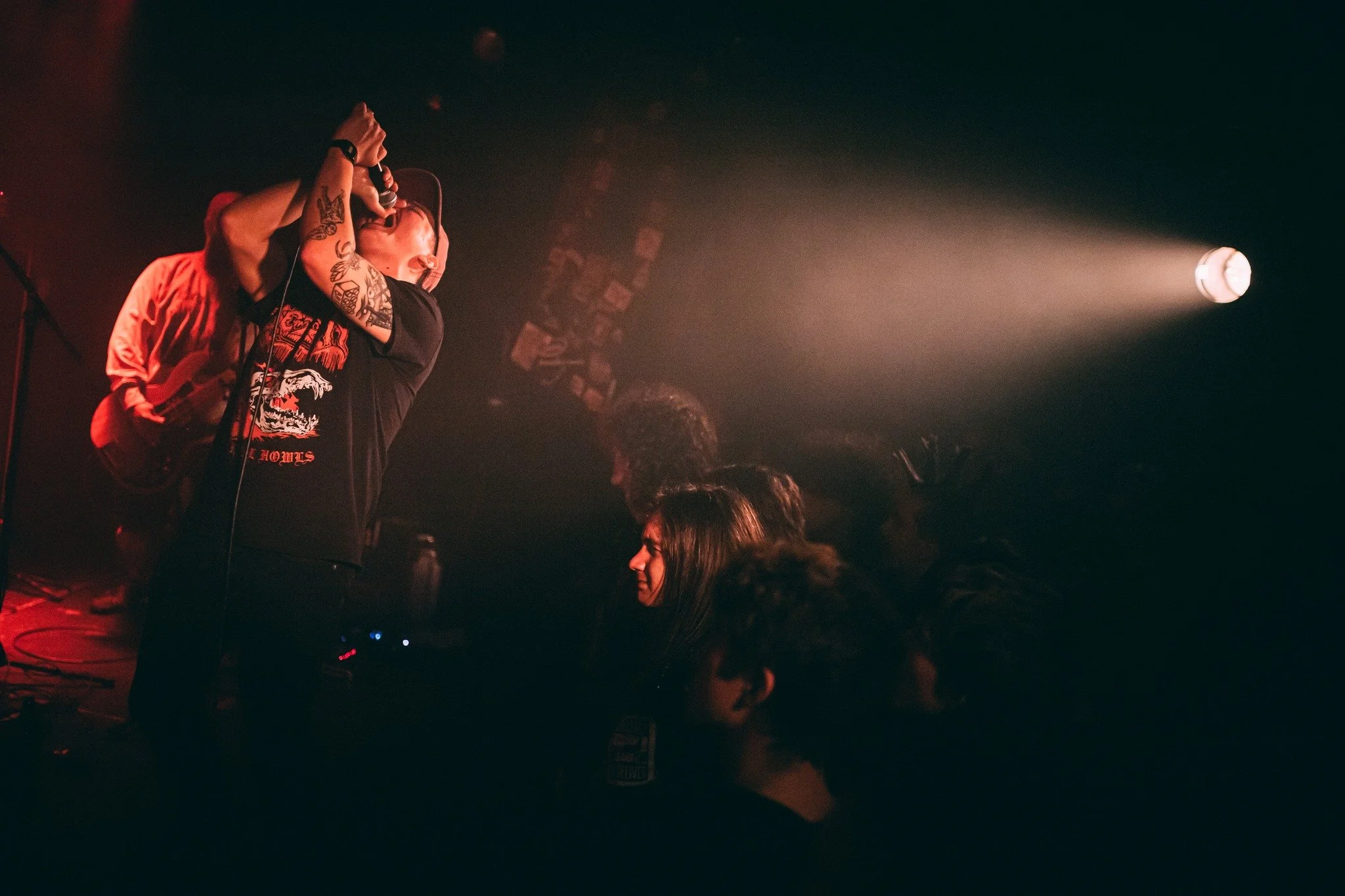 A female singer with short hair, tattoos on her arms, wearing a black T-shirt, passionately performing on stage with a microphone, with stage lights and audience members visible.
