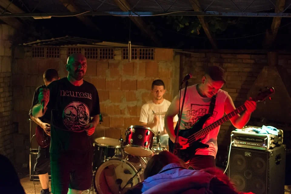Four musicians performing on stage in a dimly lit setting with brick and wooden walls, including a vocalist, drummer, and bassist.