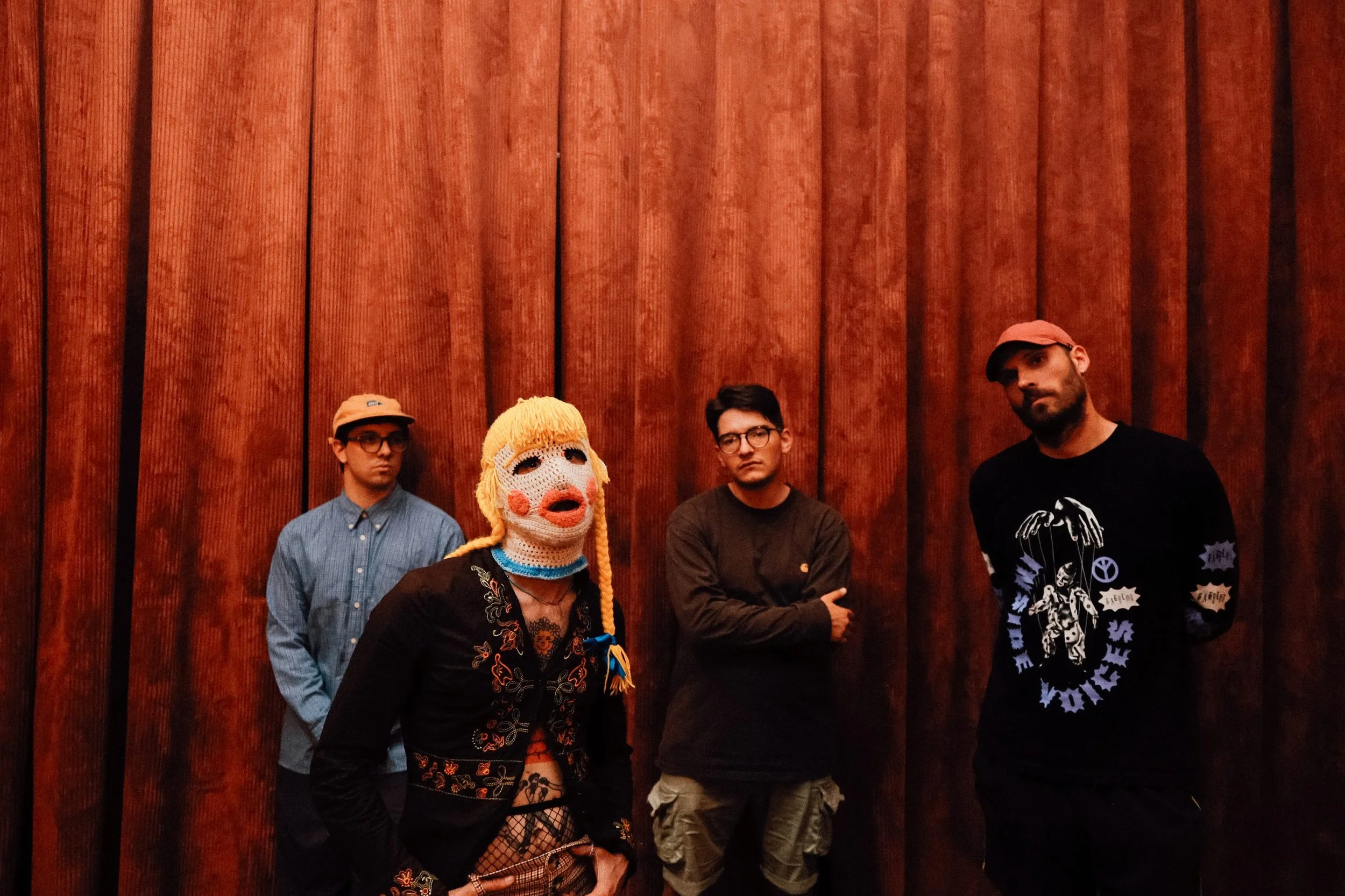 Four people standing in front of a wooden paneled wall. The person in front is wearing a knitted mask with orange hair and red lips.