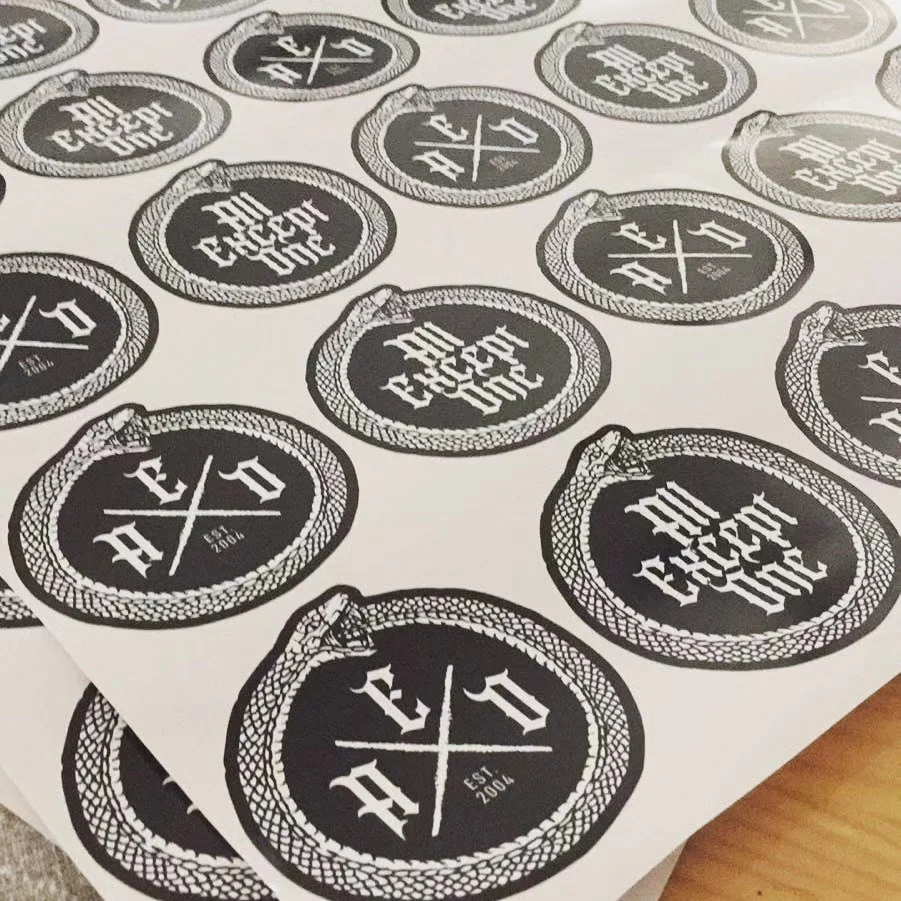 Sheet of black and white circular stickers with a logo that says 'All Except One' and 'EST 2004' inside, surrounded by a snake-like border.