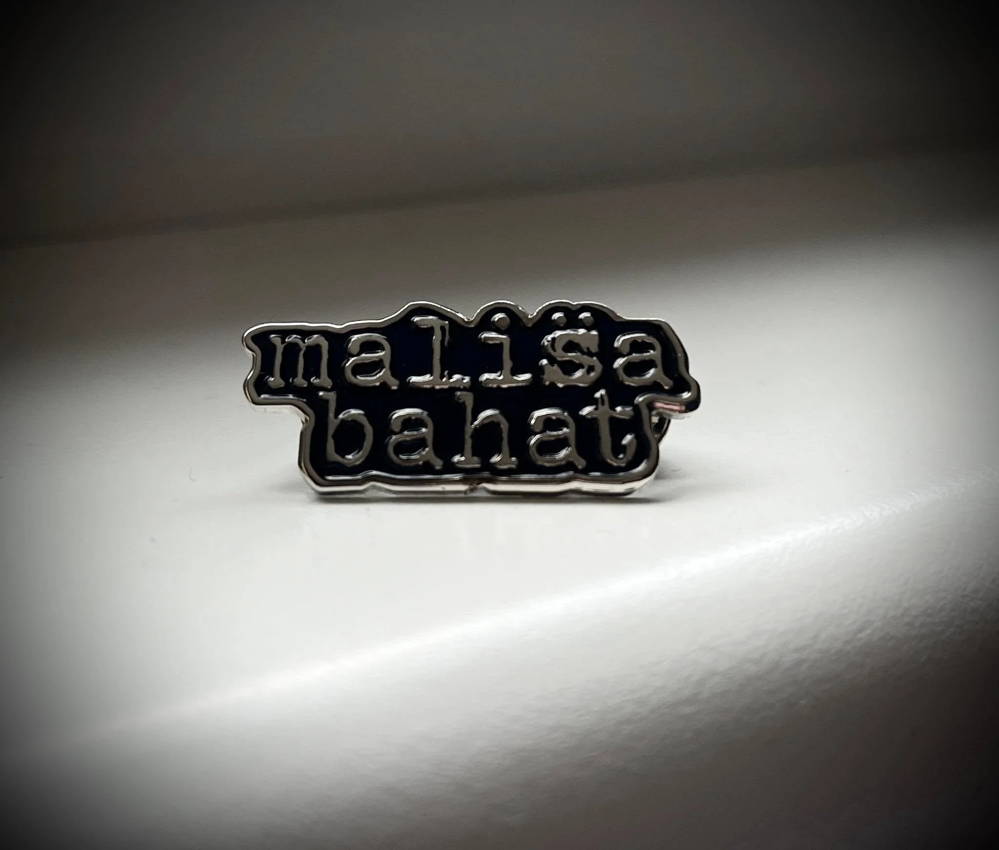 A metallic pin with the words 'Malisa' and 'Bahat' in stylized font, lying on a white surface.
