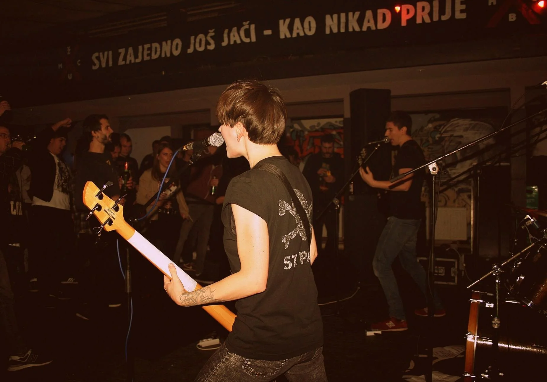 A woman with short brown hair playing an electric guitar and singing into a microphone during a live performance in a crowded venue. The background features a large banner in a foreign language.