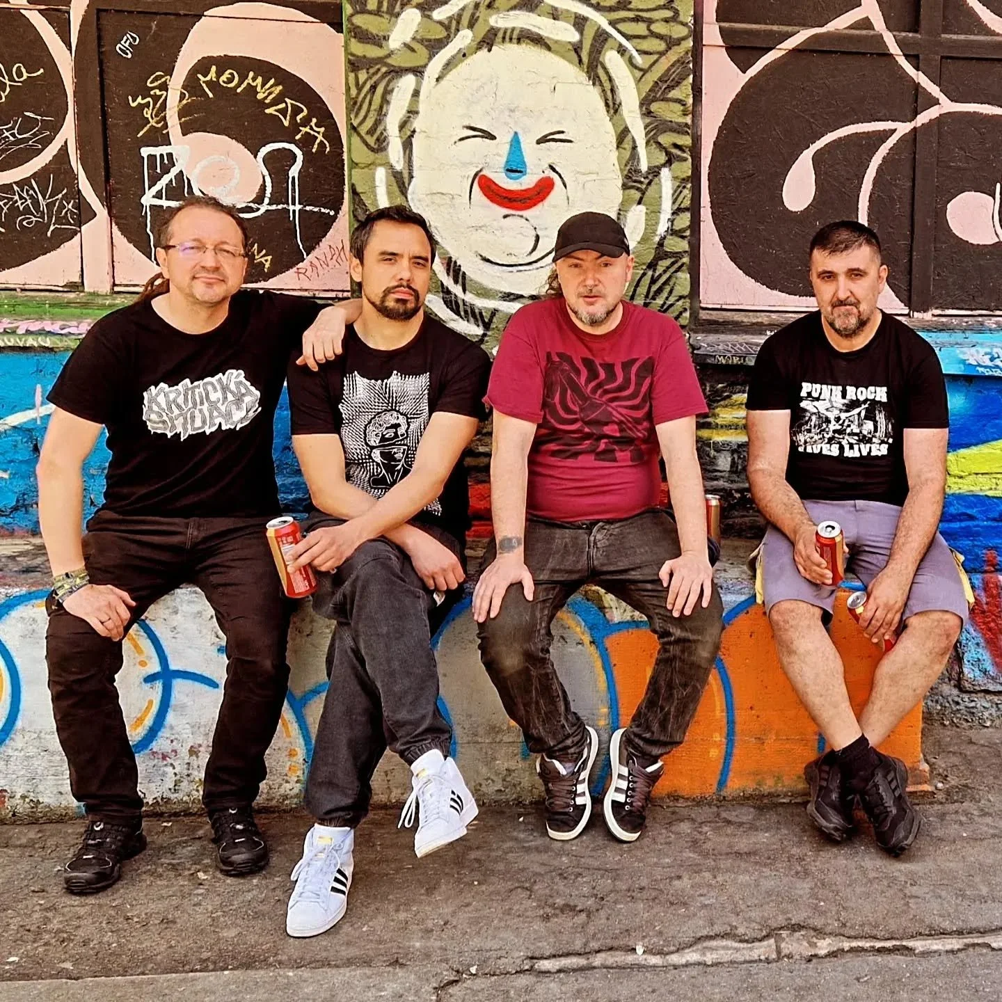Four men from band LOS PRIMJER sitting on a colorful graffiti-covered wall, holding cans of soda, with a mural of a smiling clown in the background.