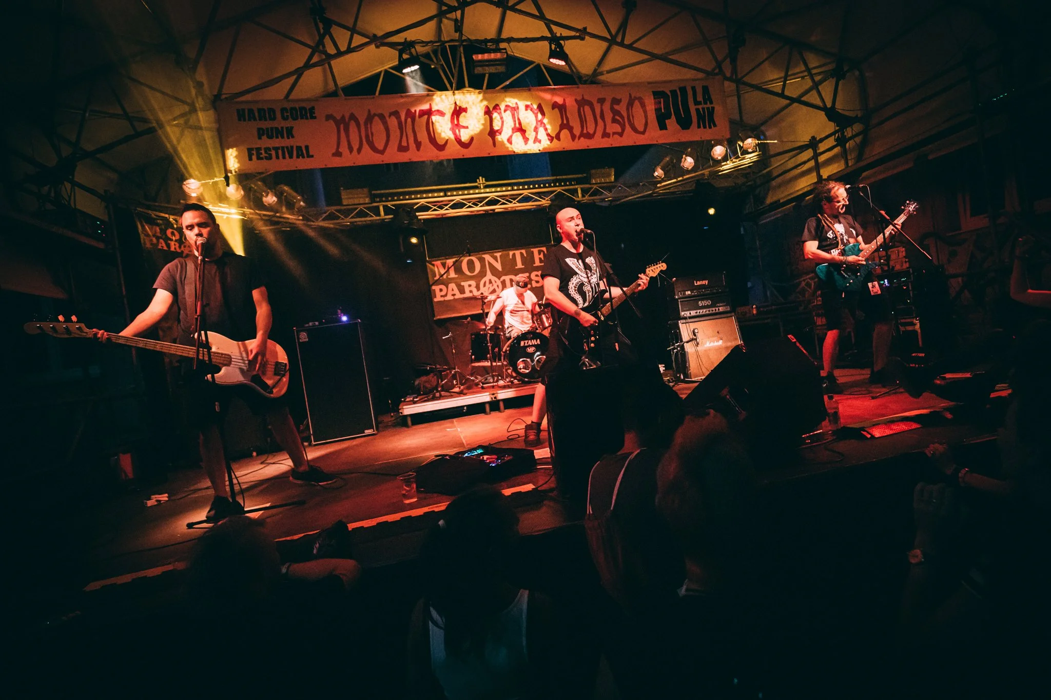 A band performs on stage at the Monte Parades Punk Festival, with four band members visible. Two guitarists and a bassist are playing, while a drummer is in the background. The stage is lit with warm lighting, and a large banner reading 'Monte Parade