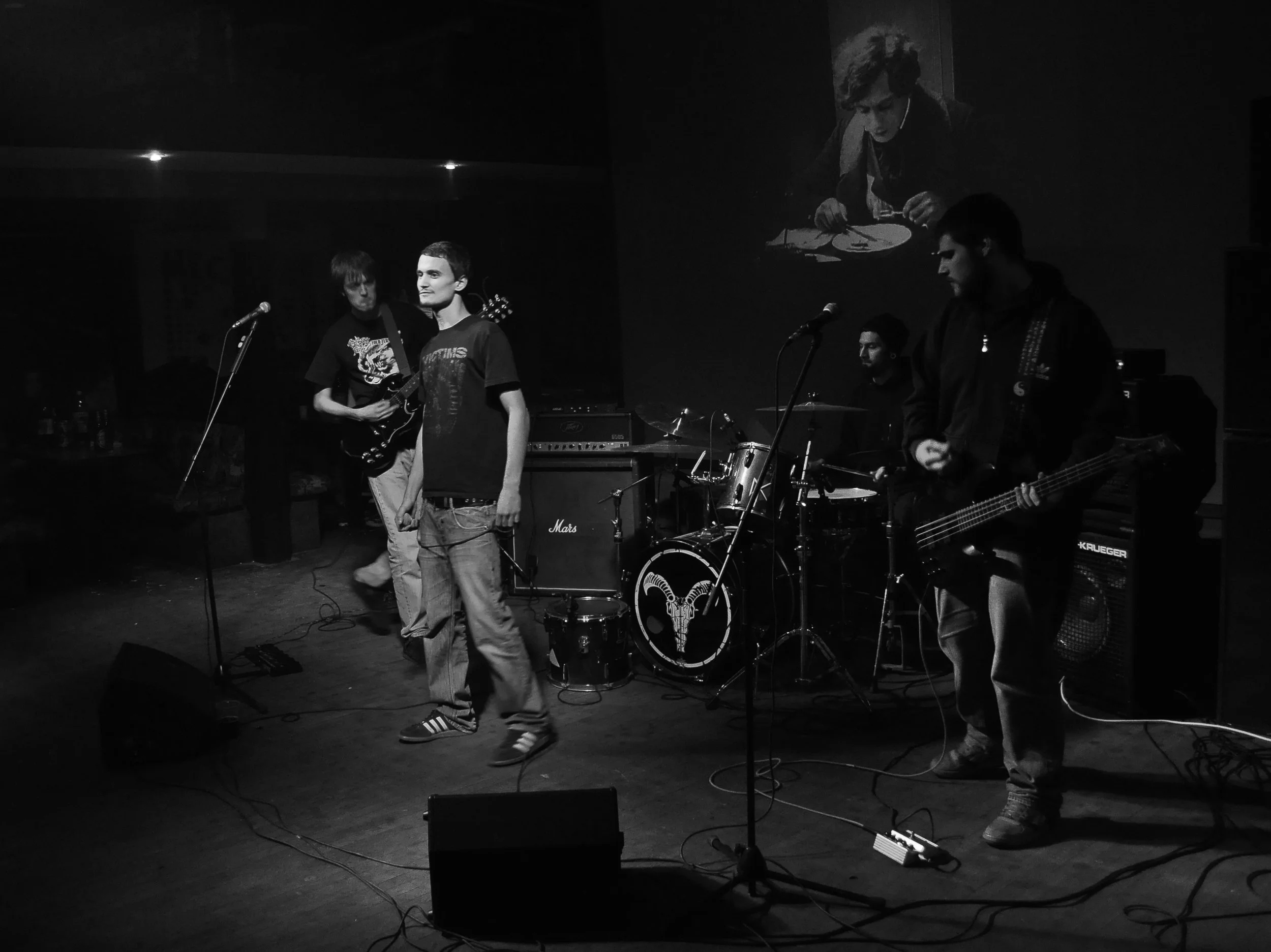 A band of four young men performing on stage with musical instruments, including guitars and drums, in a dimly lit indoor venue.