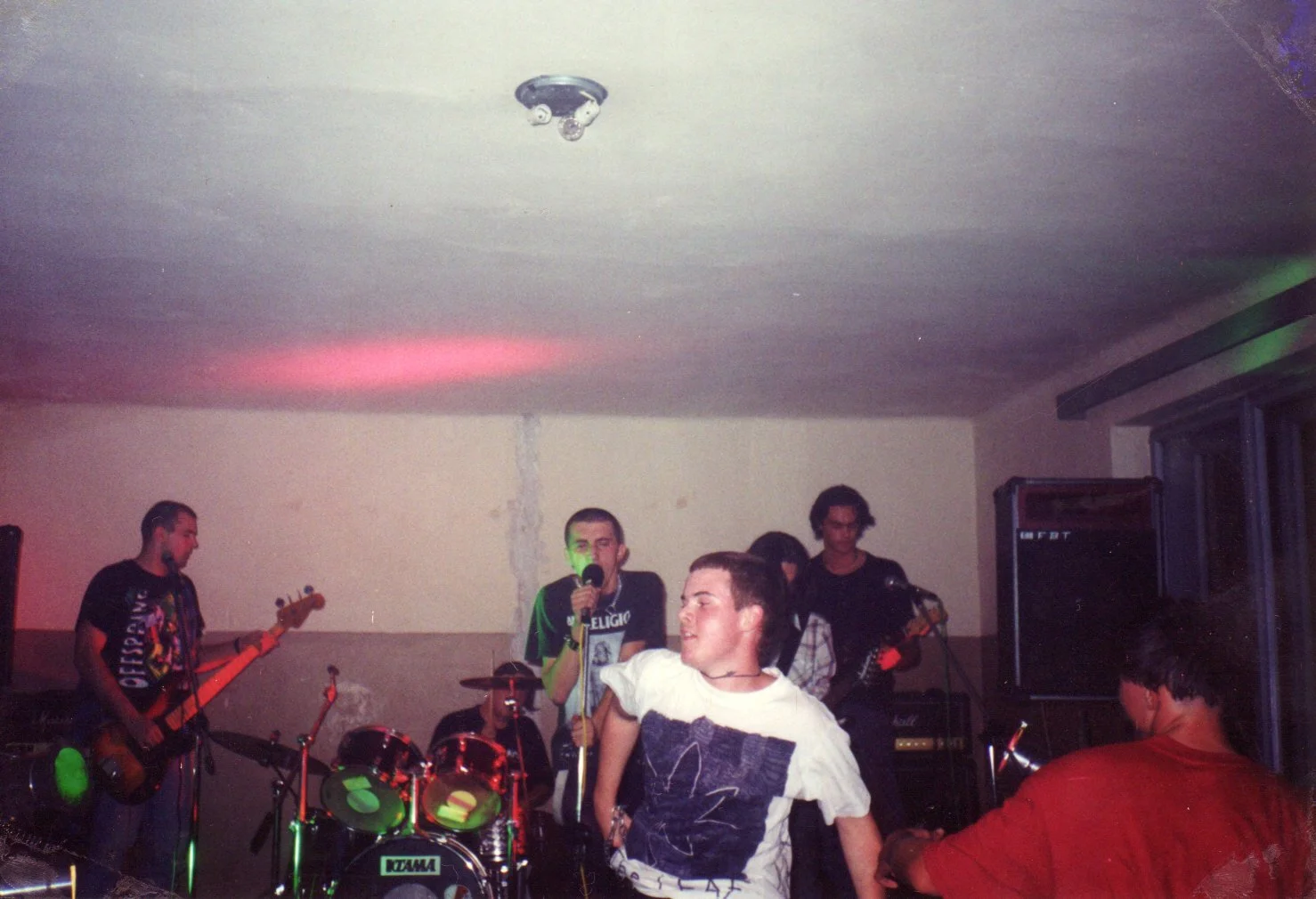 Young people performing music in a small indoor venue with a band setup, including guitars, a drum set, and microphones.