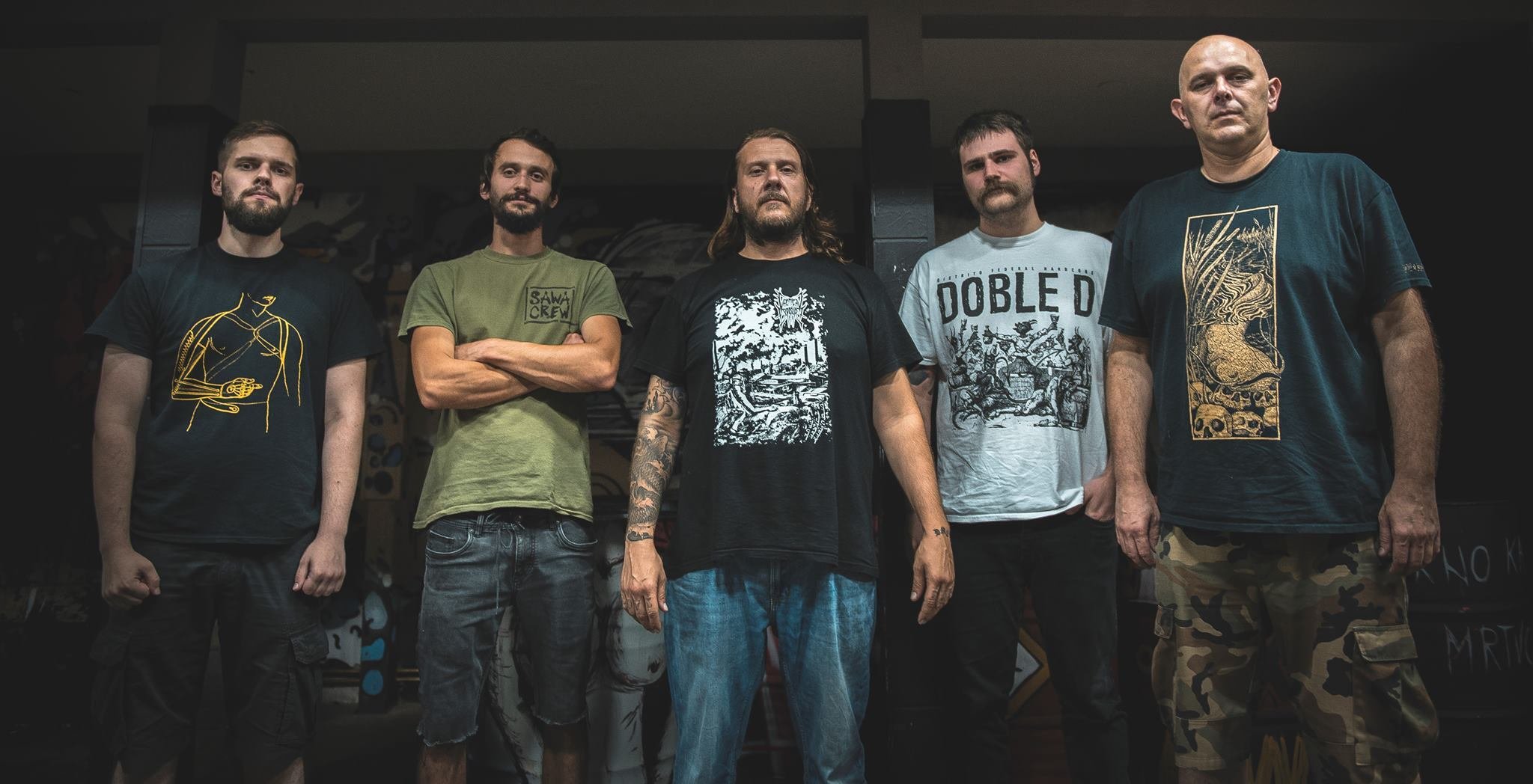 Five men standing side by side in a dimly lit indoor space with graffiti-covered walls in the background.