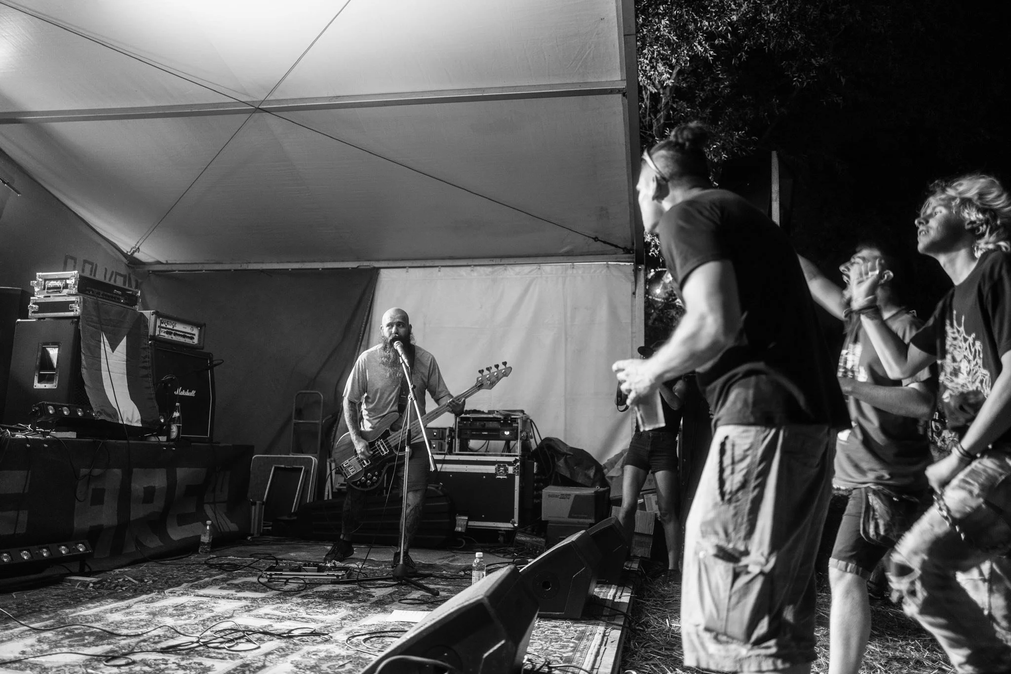Black and white photo of a small outdoor concert with a band performing on stage and an audience watching. The band includes a bearded man playing a bass guitar and a woman singing or speaking into a microphone. The stage is covered and has various m