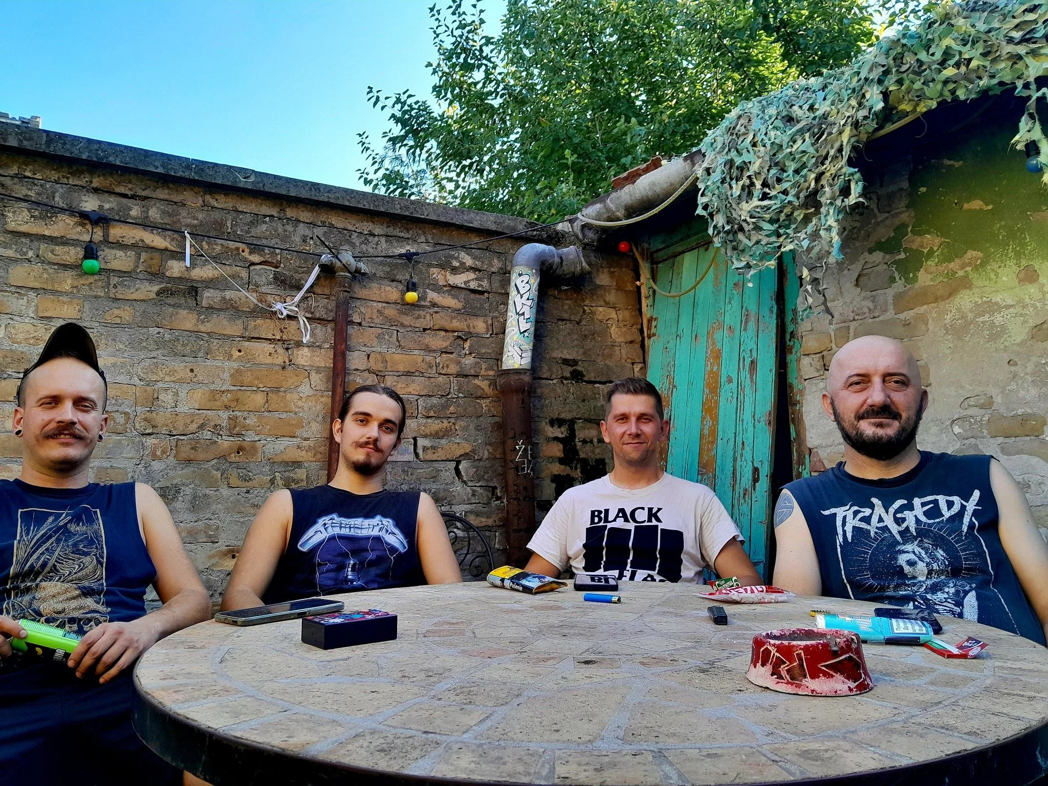 Group of five men sitting at an outdoor stone table, smiling and looking at the camera, with a rustic brick wall, green door, and foliage in the background. Items on the table include snacks, a lighter, and personal belongings.