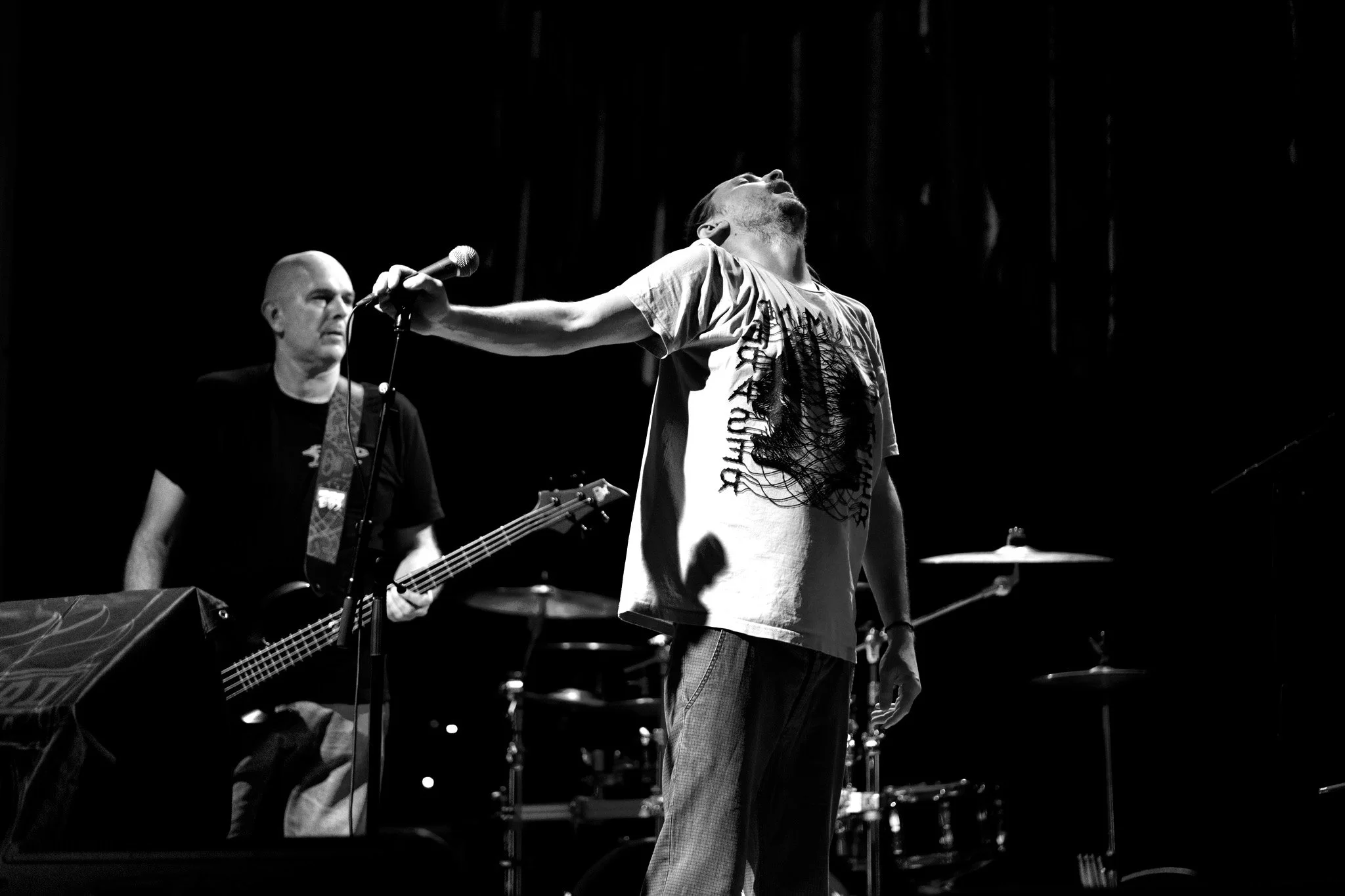 Black and white photo of a male lead singer performing on stage with his head thrown back, one hand holding a microphone and the other hand relaxed by his side, wearing a t-shirt and plaid pants. Behind him, a guitarist plays guitar while looking serious, and drums are partially visible.