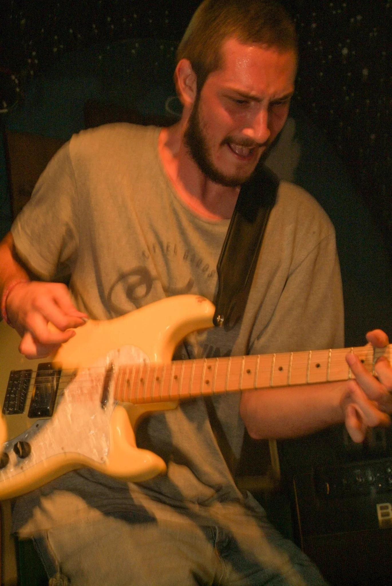 A man with short hair and a beard passionately playing an electric guitar in a dimly lit setting.
