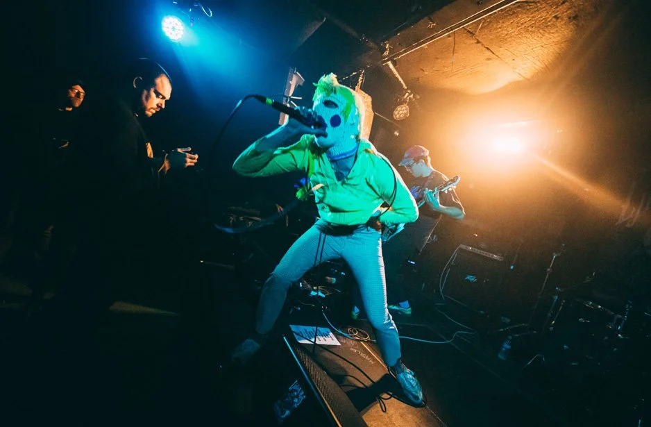 A person wearing a colorful animal mask and bright green hoodie singing into a microphone on stage with band members playing guitar in the background at a live concert.