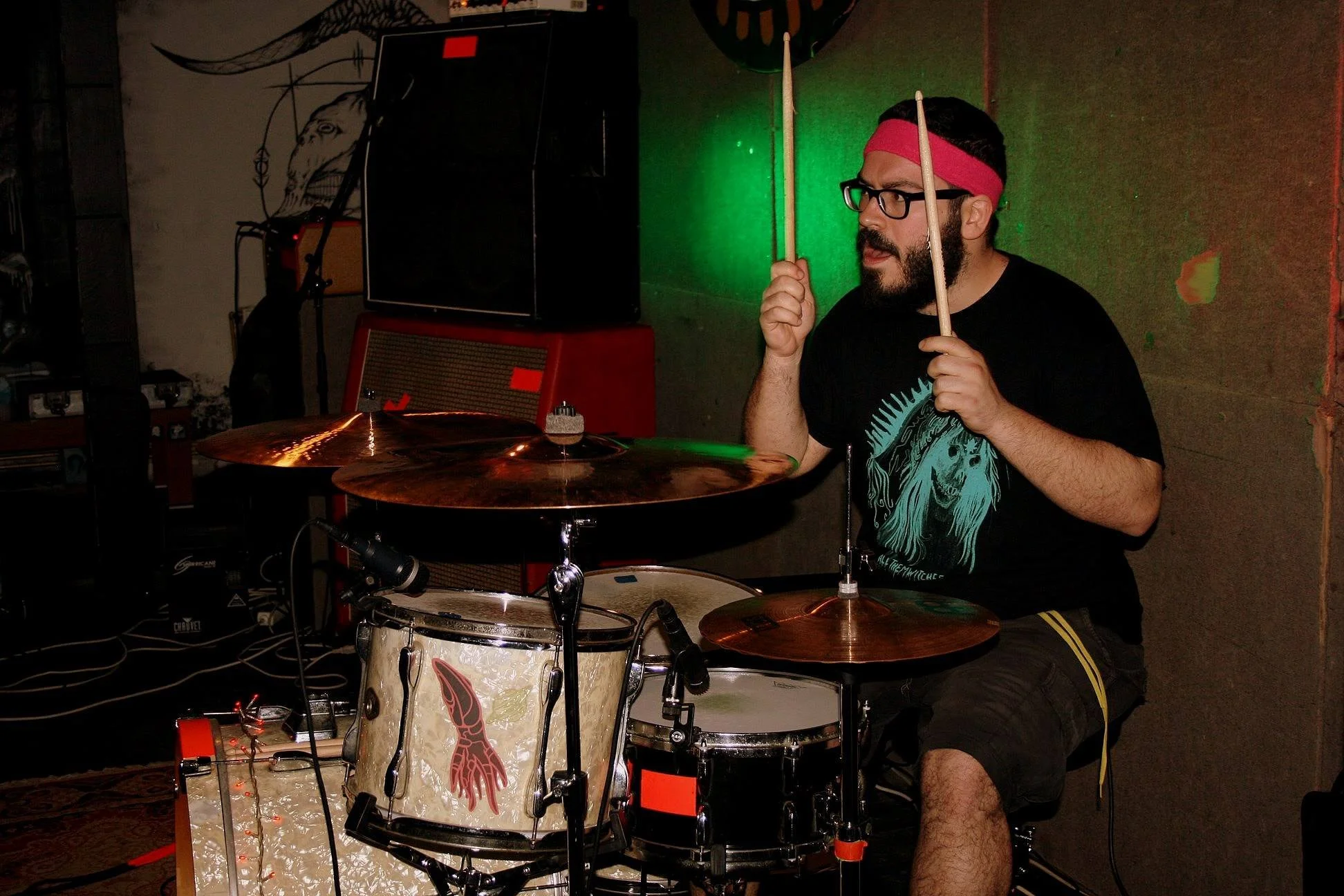 A man with glasses, a red headband, and a beard is playing drums with drumsticks. He is seated behind a drum set in a room with dark walls; there is a wall decoration and some electronic equipment in the background.