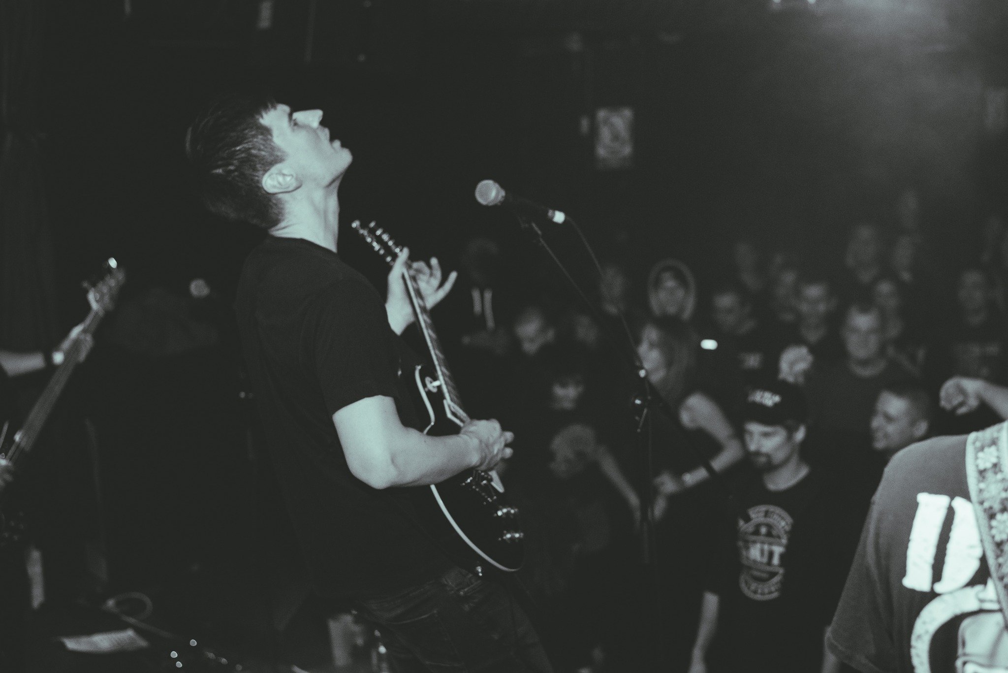 A person with short dark hair, wearing a black shirt, is performing on stage with eyes closed and head tilted up, holding a guitar, surrounded by a crowd in a dark setting.