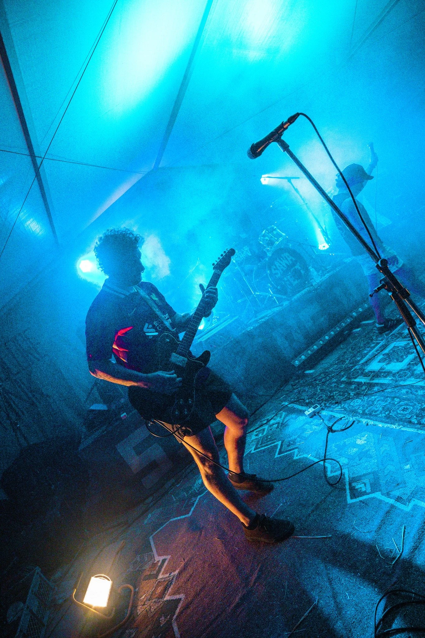 A musician playing an electric guitar on a dimly lit stage with blue lighting, microphone stands.