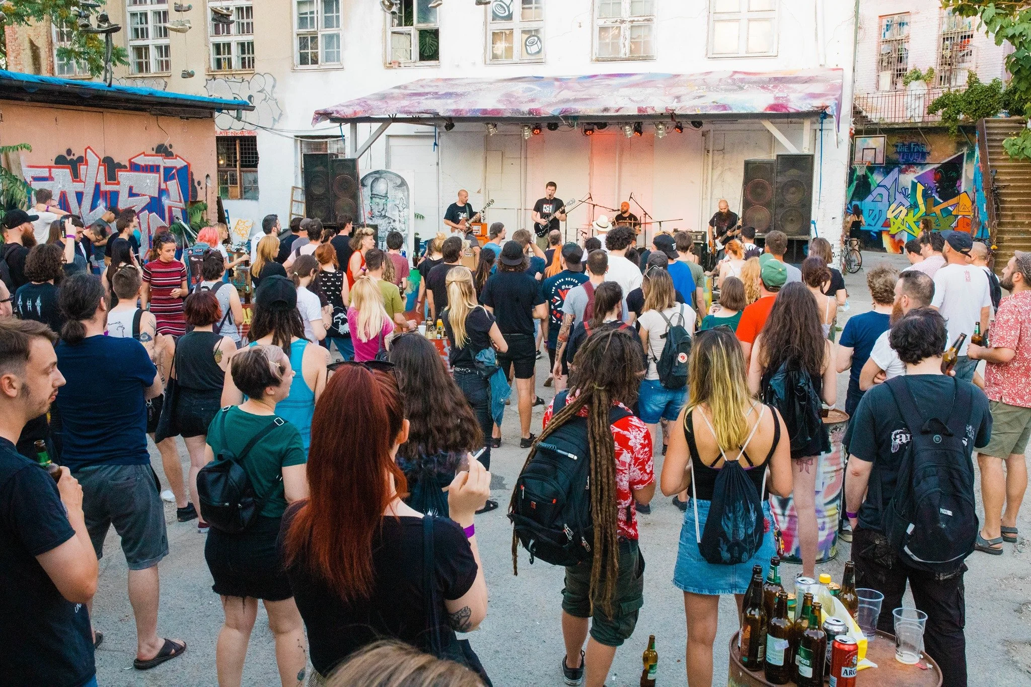 A crowd of people gathered outdoors watching a band perform on a small stage with graffiti-covered walls surrounding them. There are bottles and cups on a table in the foreground.