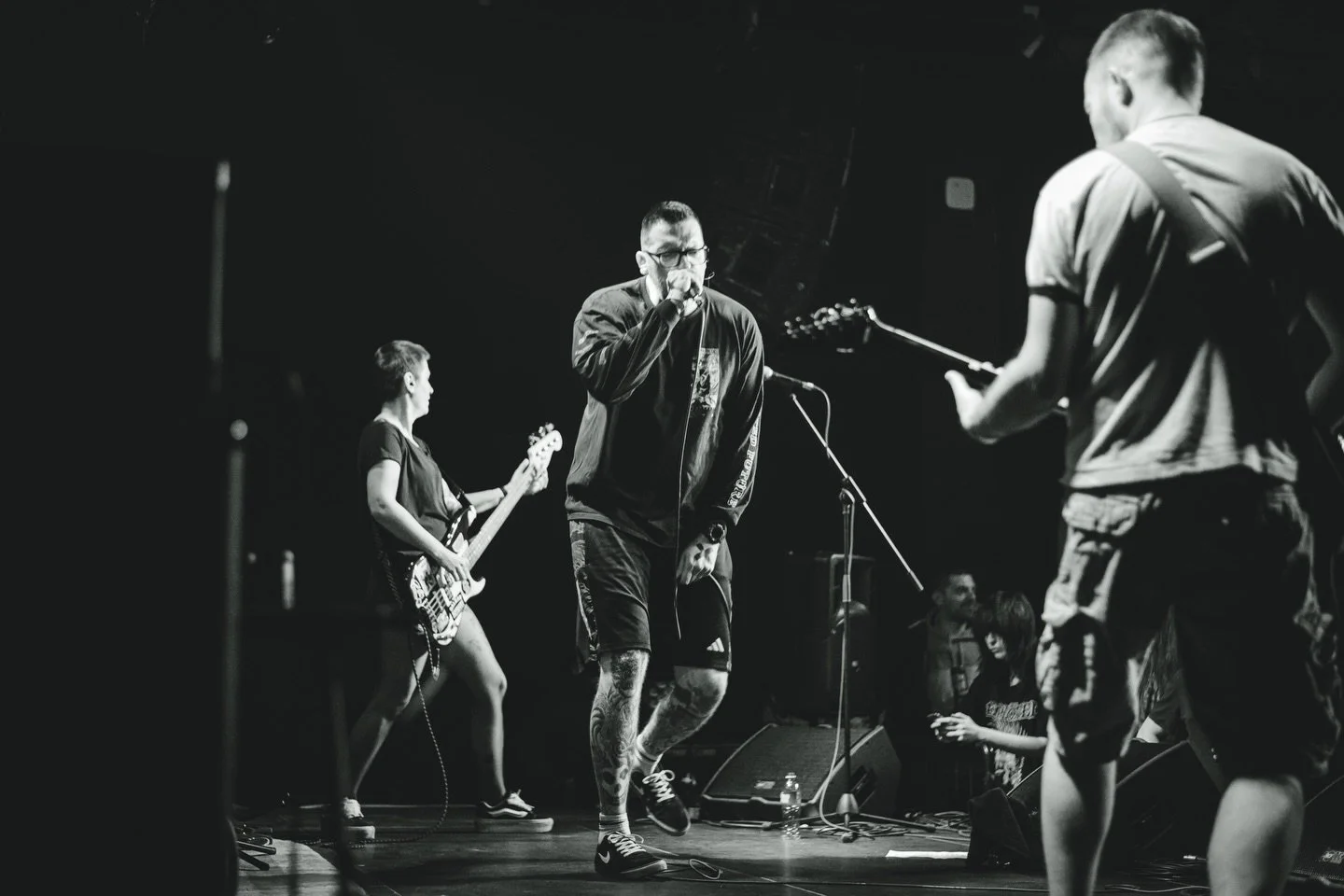 A band performs live on stage with a vocalist, guitarist, and bassist in black and white photography.