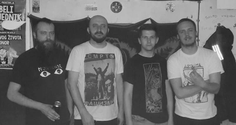 Four men standing indoors at a social gathering or event, with a dark backdrop and posters on the wall.