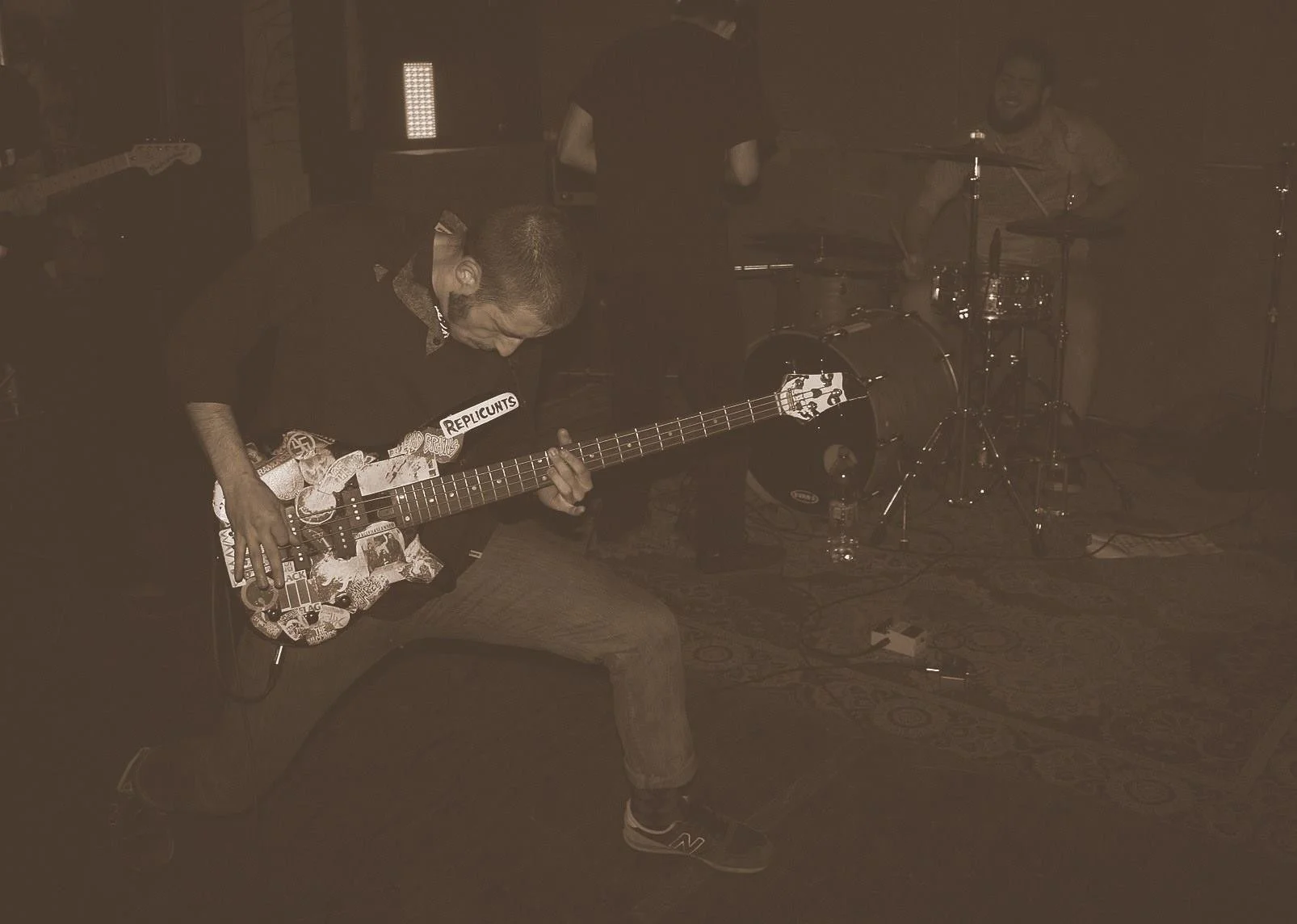 A musician kneeling and playing an electric guitar on stage, with a drummer smiling in the background, in a dimly lit environment.