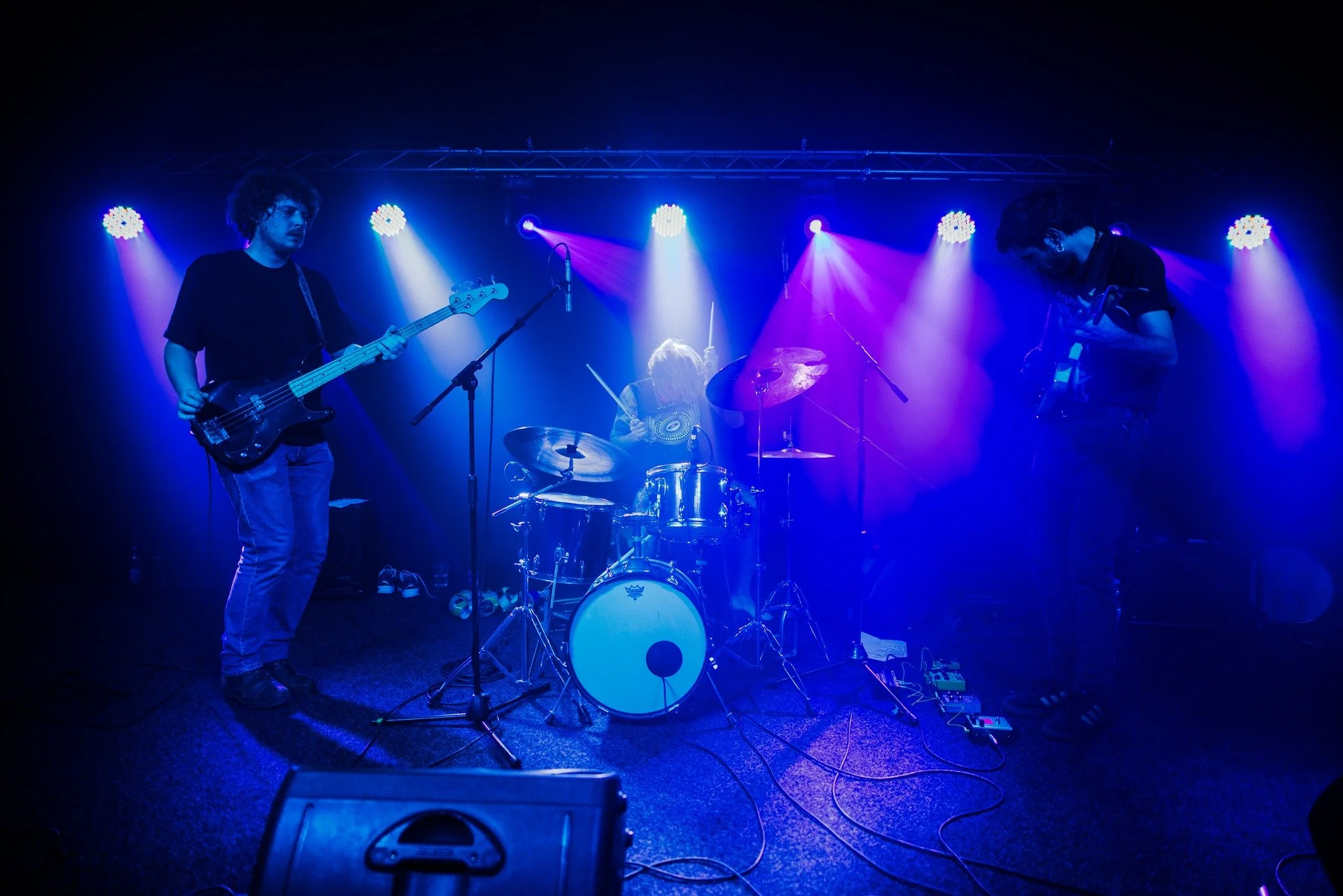 A band of three musicians performing on stage under colorful lighting. One playing bass guitar, another on drums, and the third on electric guitar.