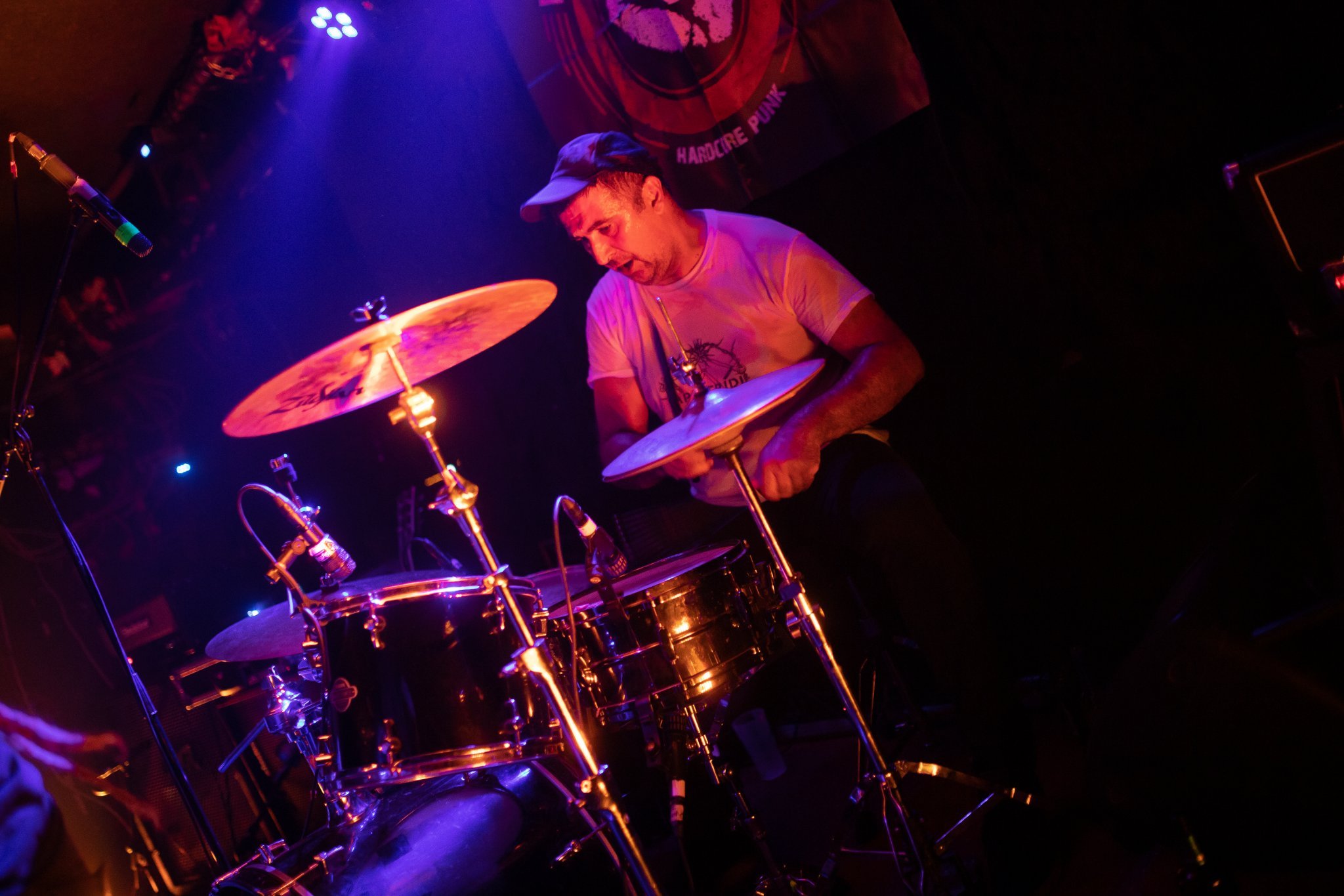 A man wearing a baseball cap playing drums on stage under purple and orange lighting.