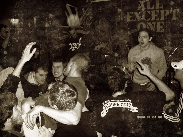 A lively group of young people dancing and singing in a bar or club with a live band performing in the background.