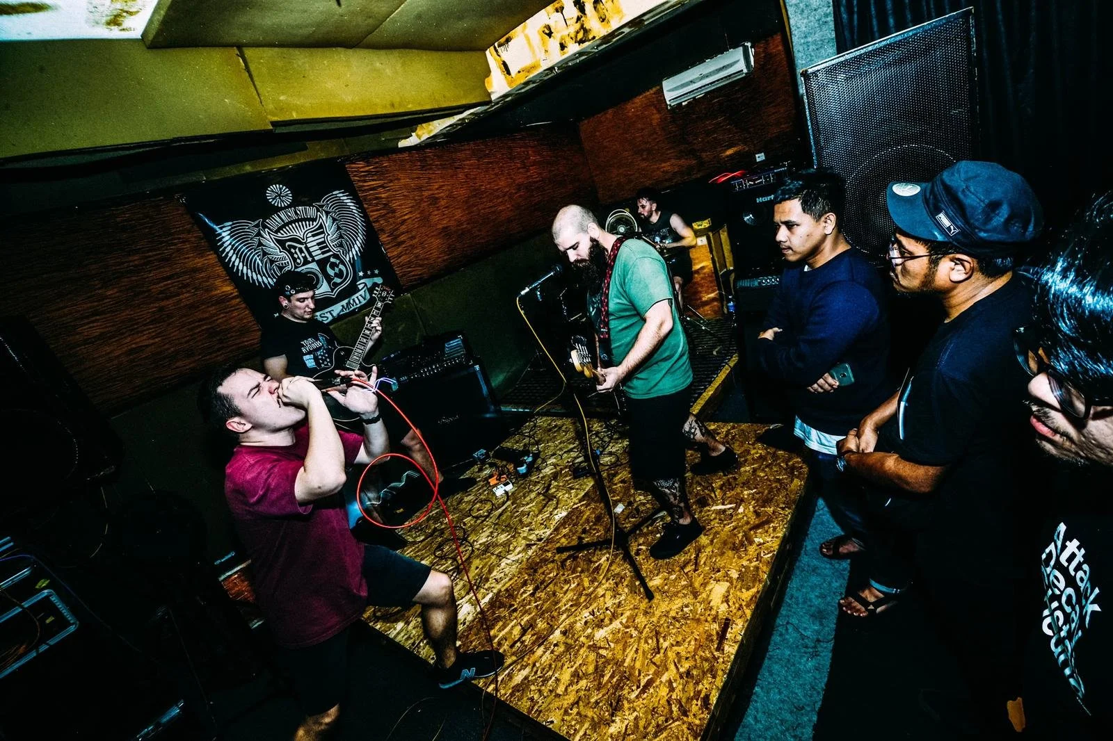 A band performs on a small stage with an audience watching closely. The drummer is singing into a microphone, guitarists are playing, a man in a green shirt is at the keyboard, and people are listening in the foreground.