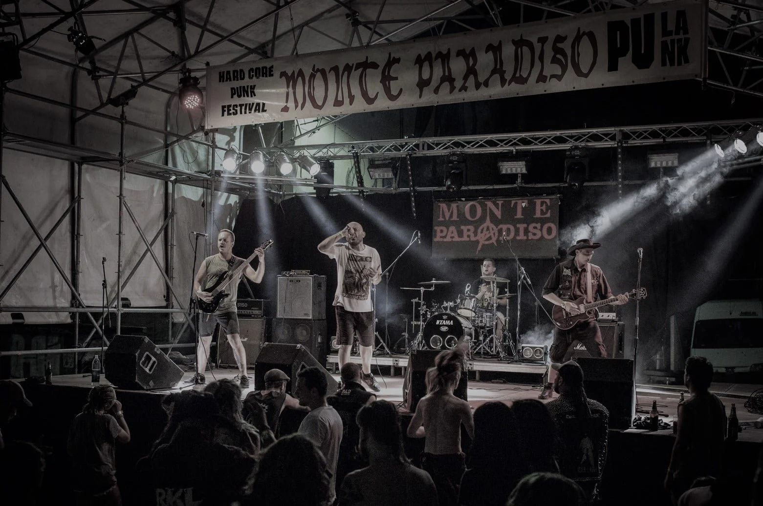 A band performs on stage at the Monte Paradiso Hard Core Punk Festival. The stage has four musicians with instruments, with a drummer in the background, and images of an audience watching.
