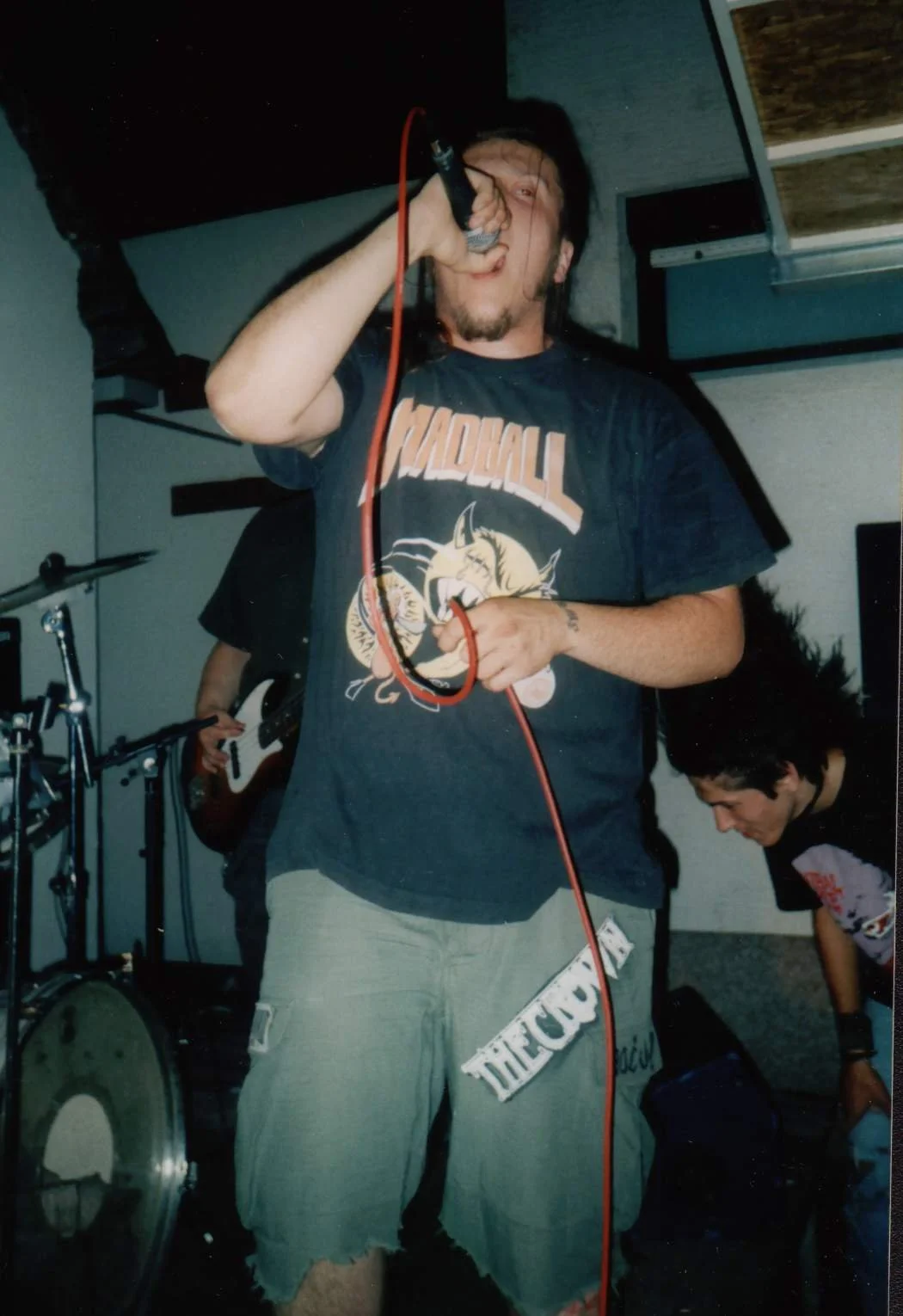 A person singing into a microphone in a band practice room, wearing a dark graphic T-shirt and torn shorts, with band members and musical instruments visible in the background.