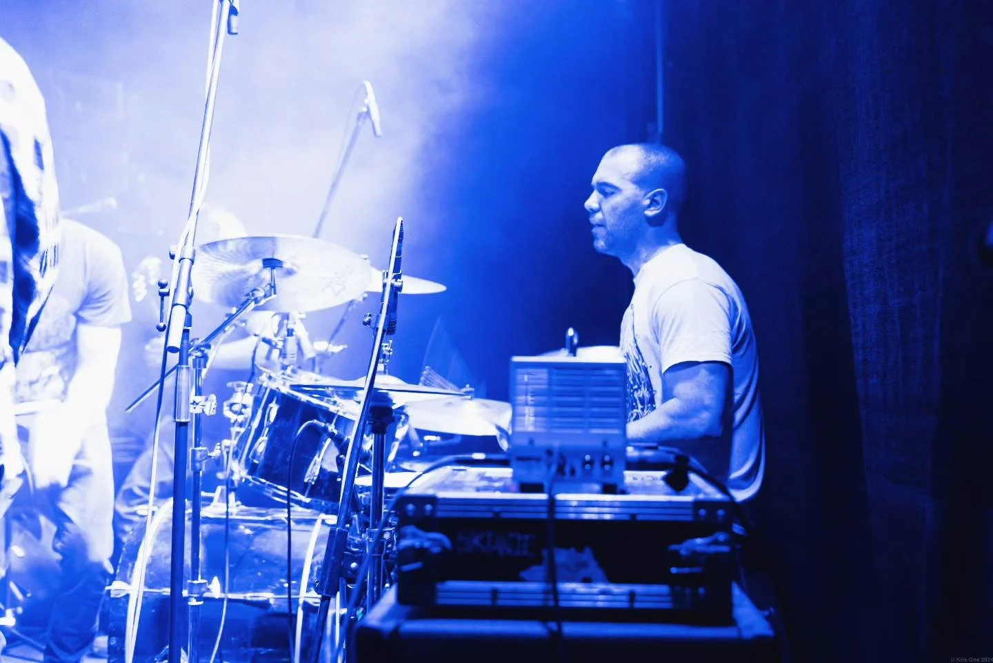 A man playing electronic keyboard on stage, illuminated in blue light with drums nearby, in a performance setting.