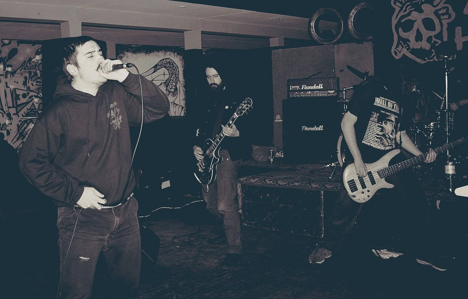 A band performing on stage with three members visible: a vocalist holding a microphone, a guitarist, and a bassist. The vocalist is wearing a dark hoodie, the guitarist has long hair and a beard, and the bassist also has long hair, all in a dimly lit
