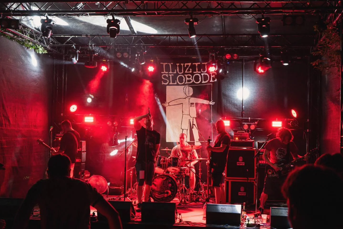 A band performing live on stage with red lighting, featuring a drum set, two guitarists, and a vocalist. A banner with a simplistic drawing of a person and the words 'ILUZIJE SLOBODE' hangs in the background.