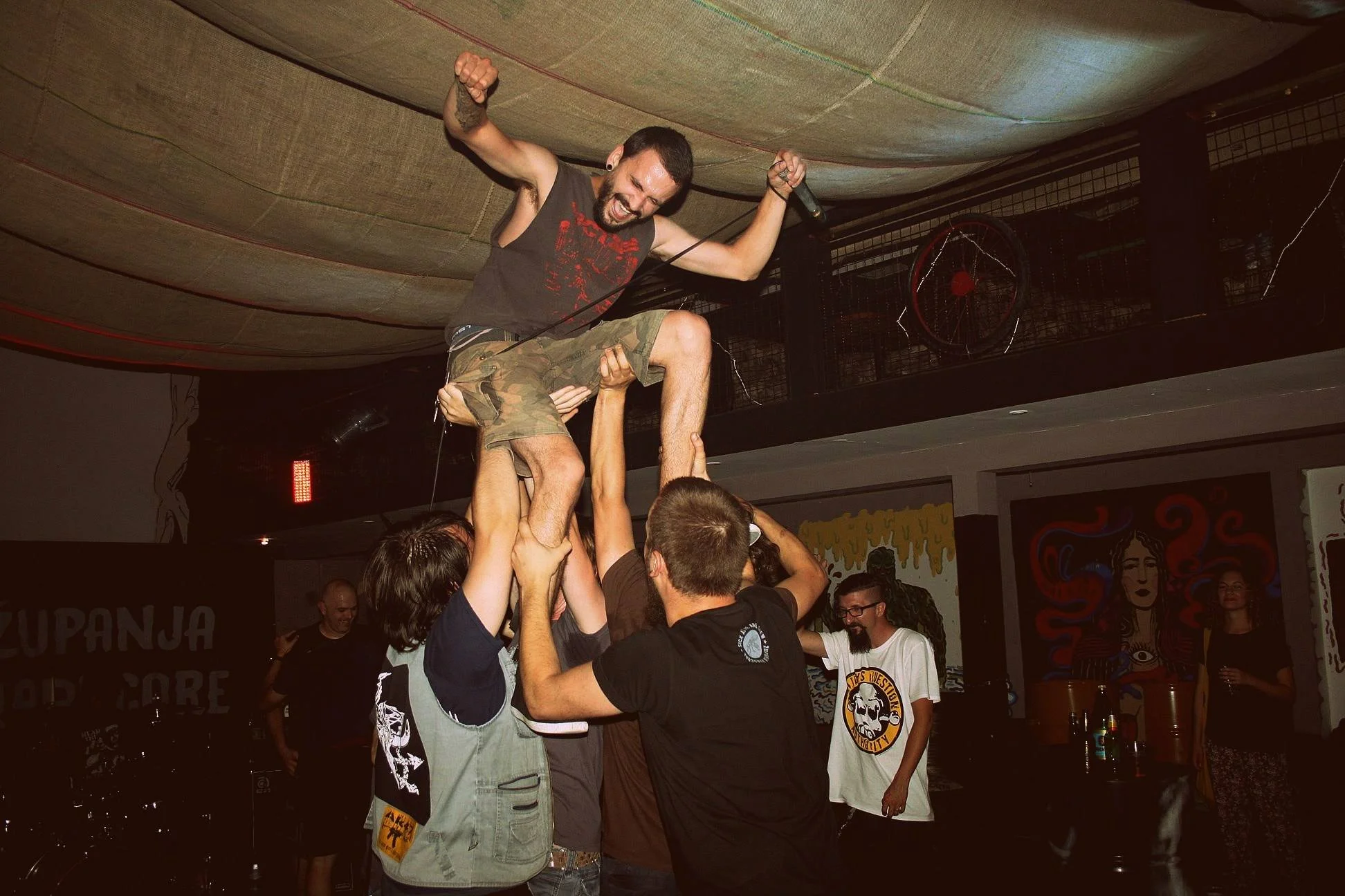 A crowd of people holding up a man with a microphone on a stage or platform during a concert or event. The man is wearing a sleeveless shirt and camo shorts, appearing to enjoy himself.