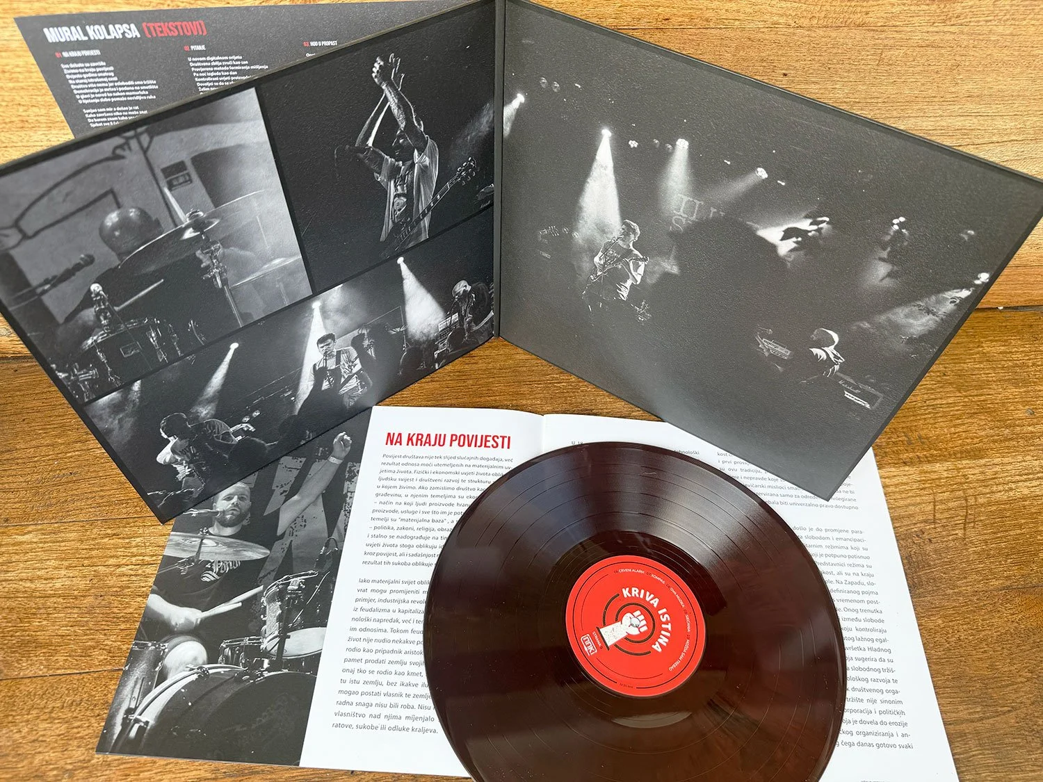 A vinyl record with red label, collectible booklet with black and white photos of musicians performing live on stage, and a black and white paper with text in a foreign language, all laid on a wooden surface.