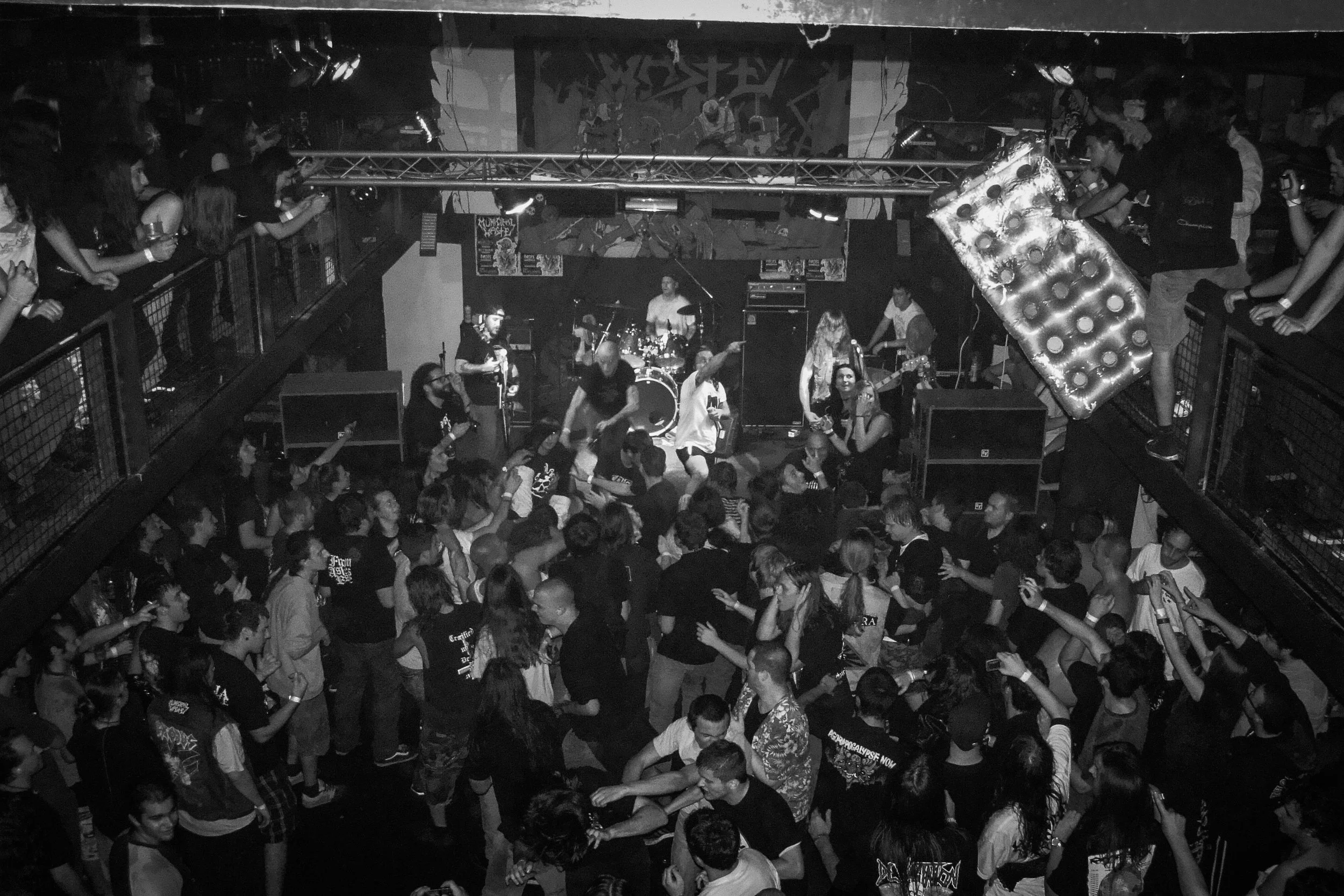A crowded indoor music concert with a band performing on stage, surrounded by audience members dancing and enjoying the show. The photo is in black and white. Band name: Municipal Waste