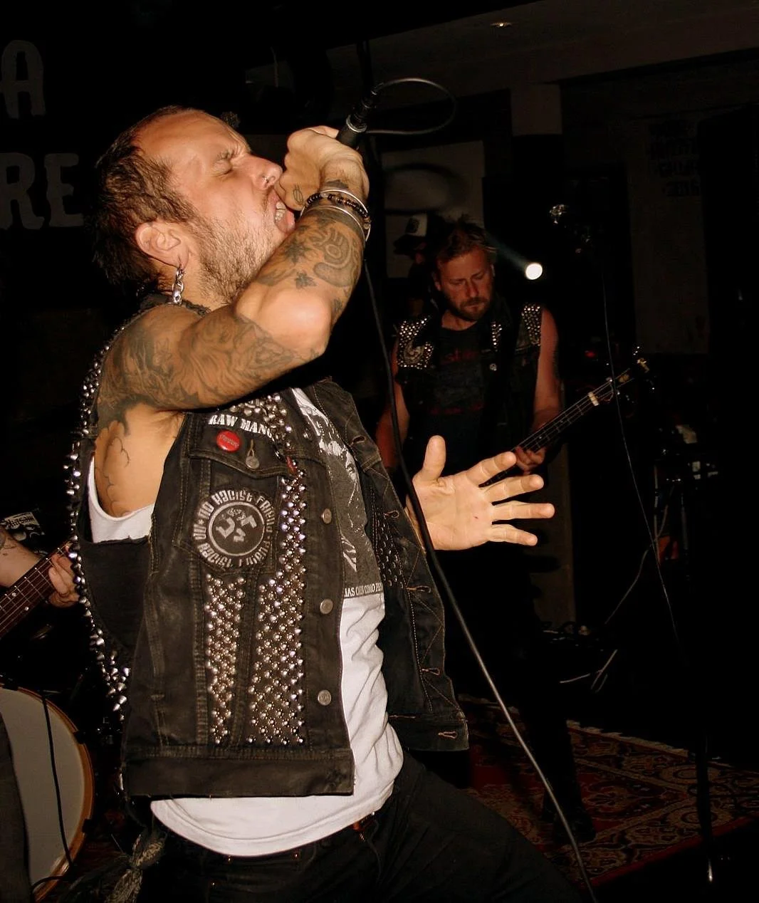 A male vocalist with tattoos singing into a microphone on stage, wearing a sleeveless vest with jewelry, with a guitarist playing in the background.