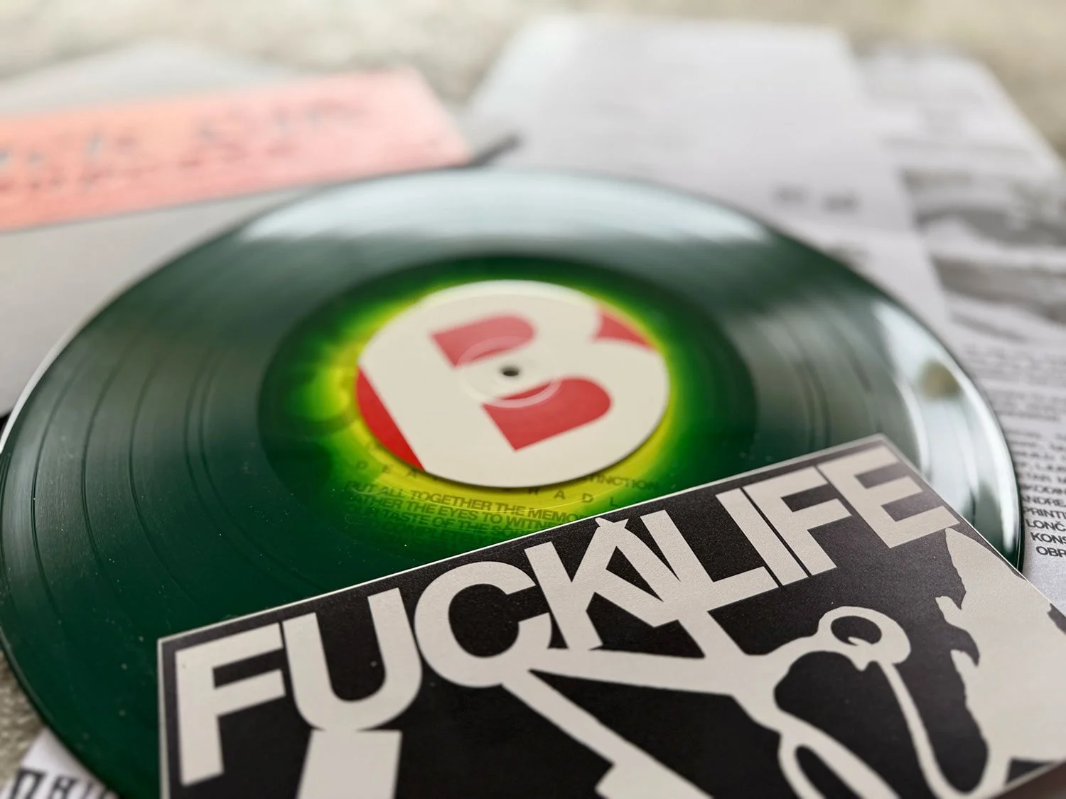 Close-up of a vinyl record with a green label and a large letter "B" in the center, partly covered by a sticker that says "FUCK LIFE" with a graphic of a person holding an object. Suplexx
