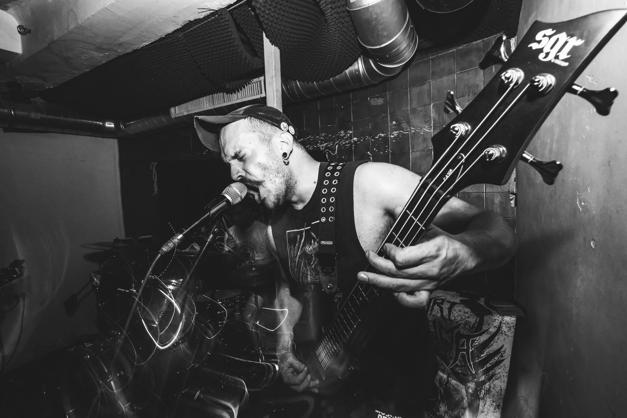 A musician with a mohawk hairstyle and piercings is singing into a microphone while playing a bass guitar in a dimly lit room with soundproofing foam and ducts on the ceiling.