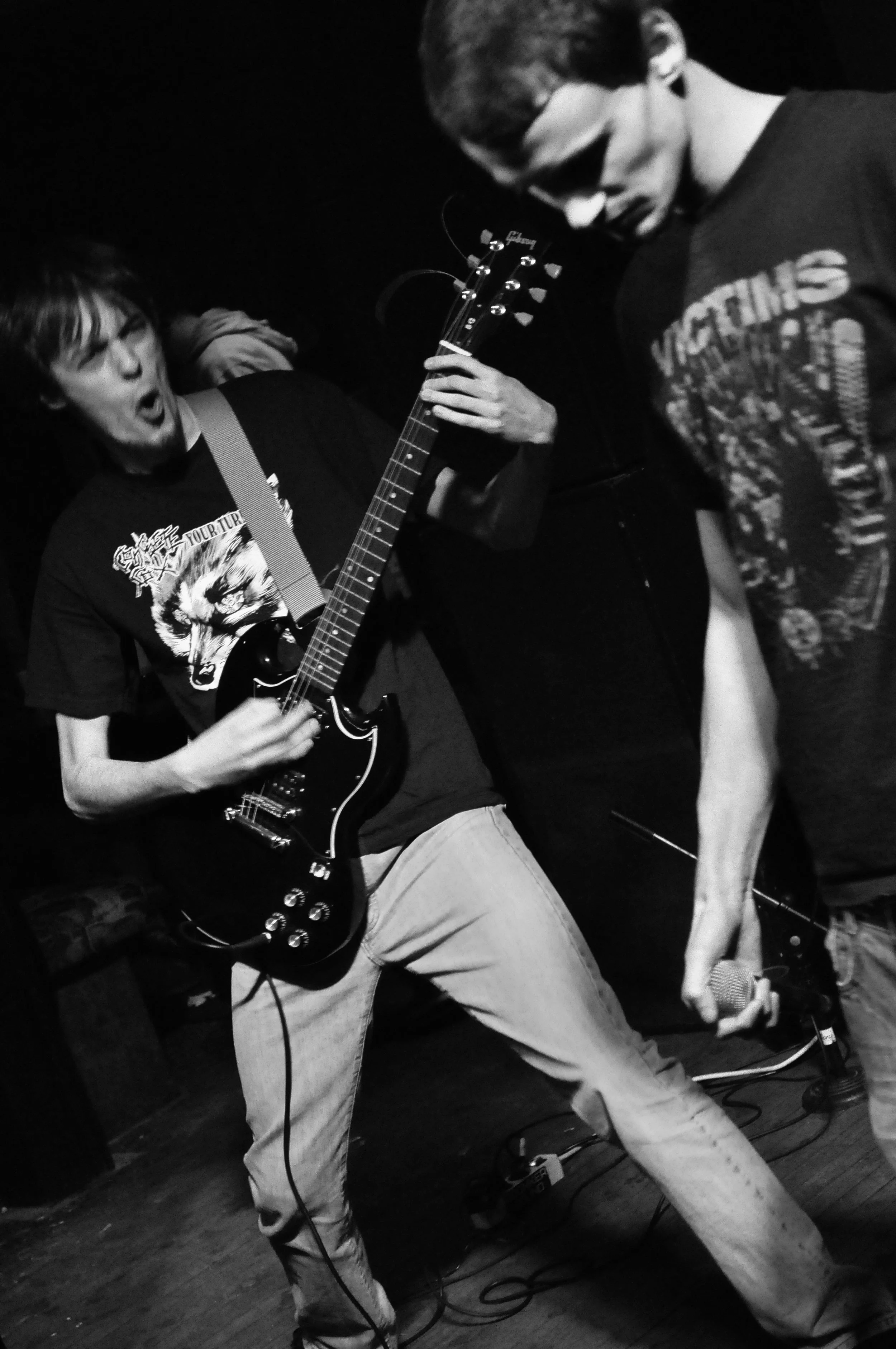Two young men playing guitar and singing passionately on stage in a dark room.