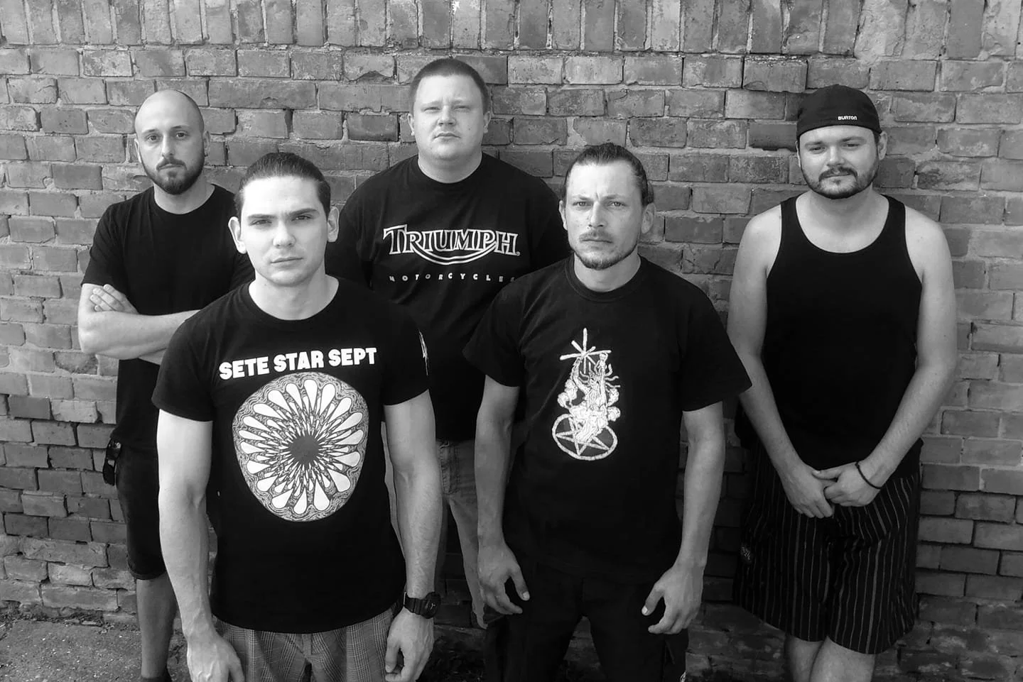 Five men standing in front of a brick wall, wearing black shirts and shorts, with serious expressions.